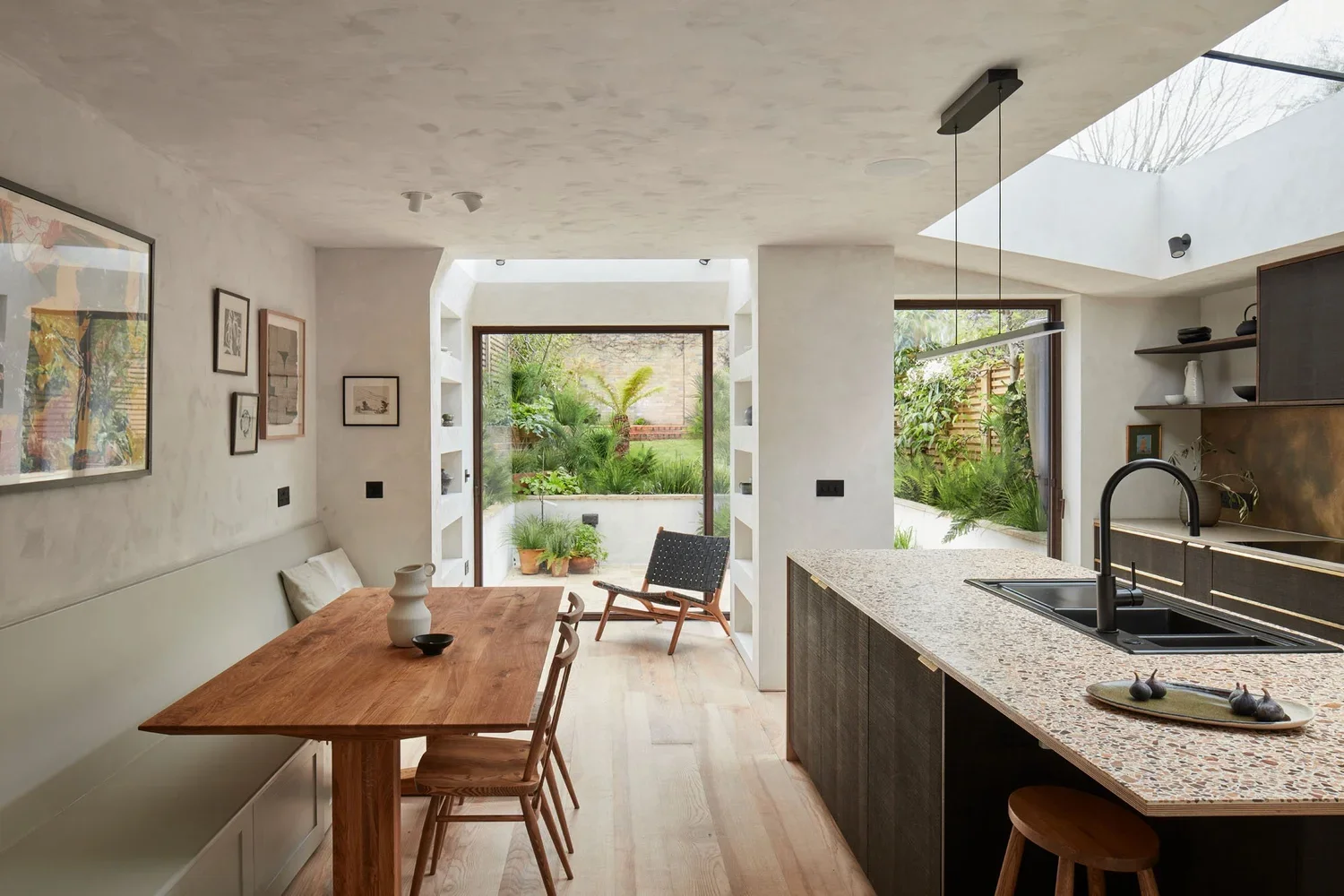 Modern kitchen and dining area with a view of a lush garden through large open windows and skylights. Features include a wooden dining table, black chairs, an island with a granite countertop, open shelving, and minimal decor.