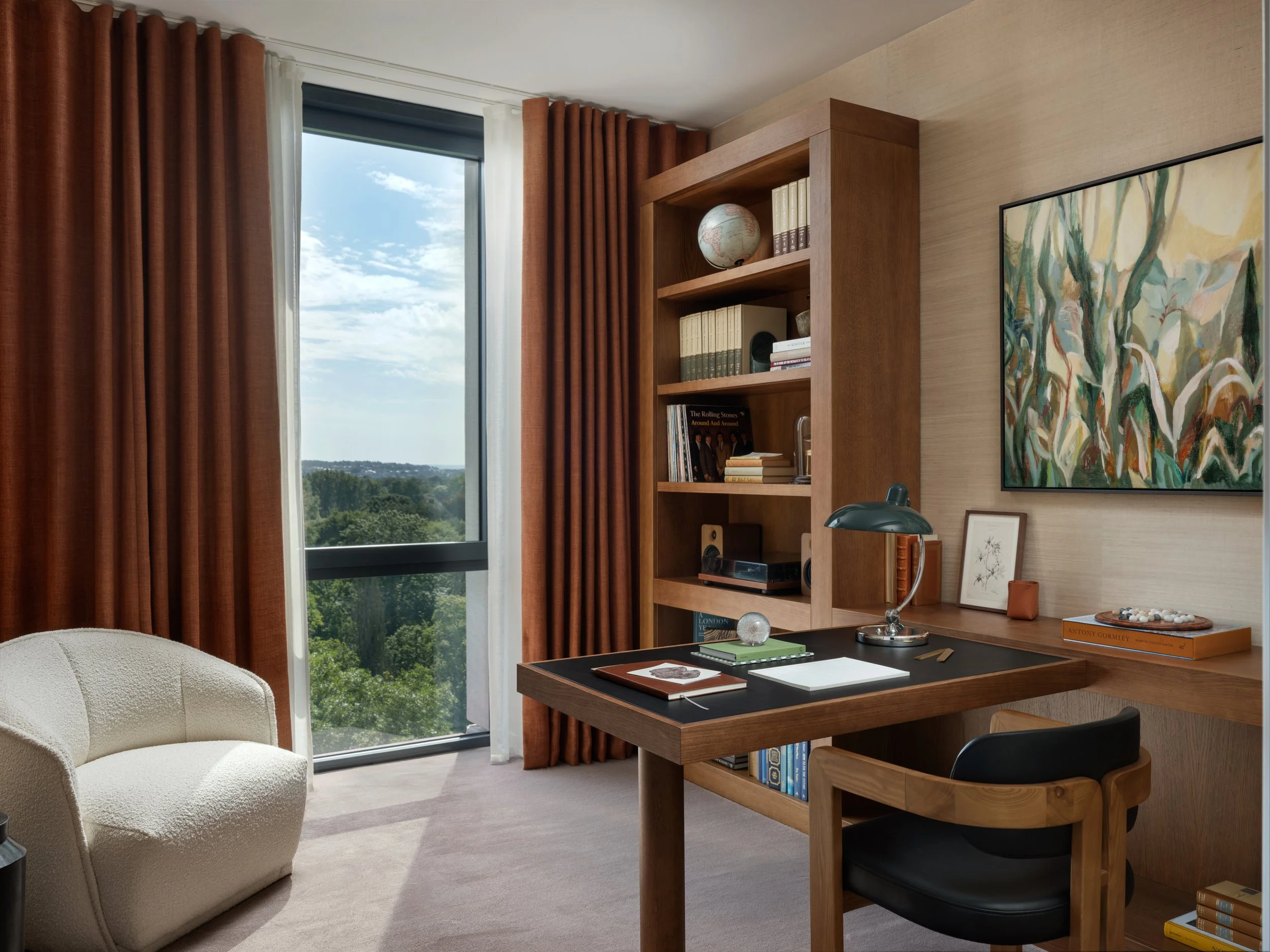 A well-lit home office with a desk, black chair, beige armchair, wooden shelves with books and decor, a large window with view of trees and sky, and abstract wall art.