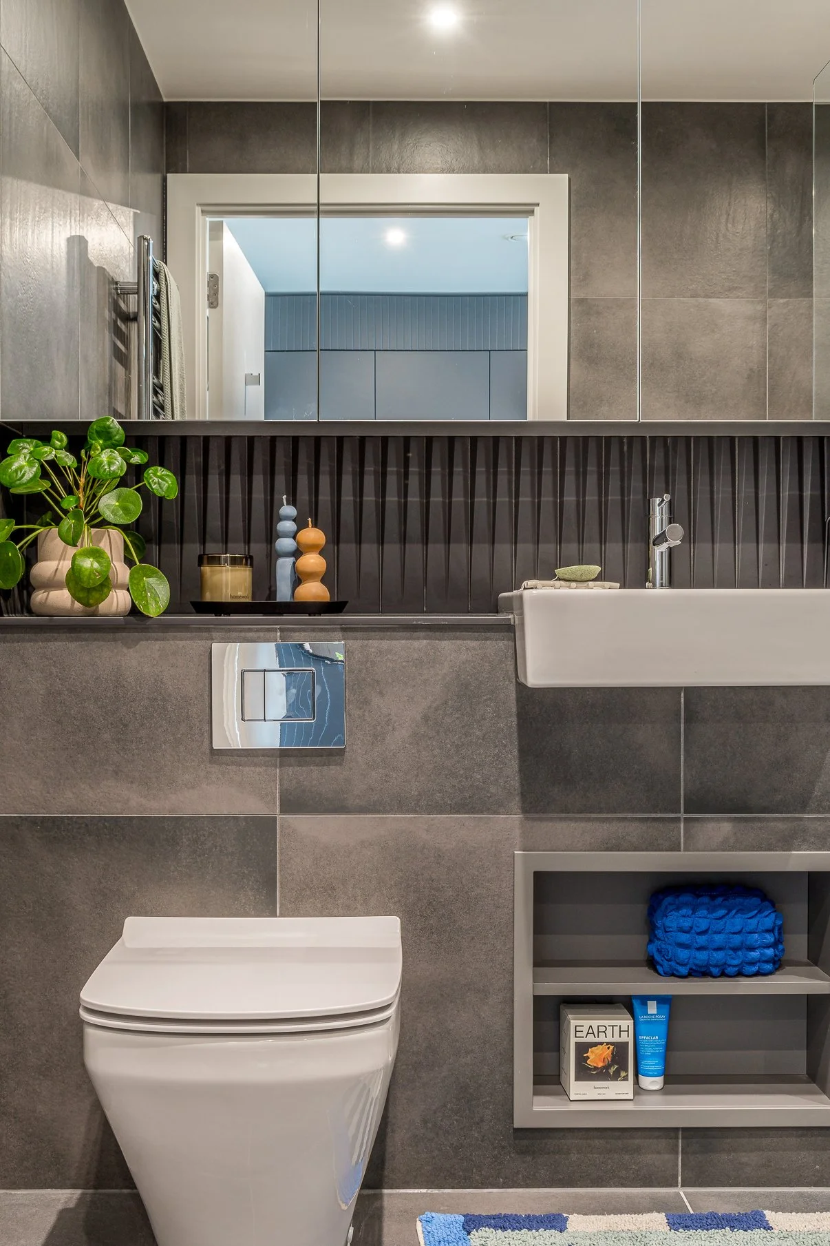 Modern bathroom with gray tiles, a white toilet, a small white sink, decorative plant, and shelf with toiletries.