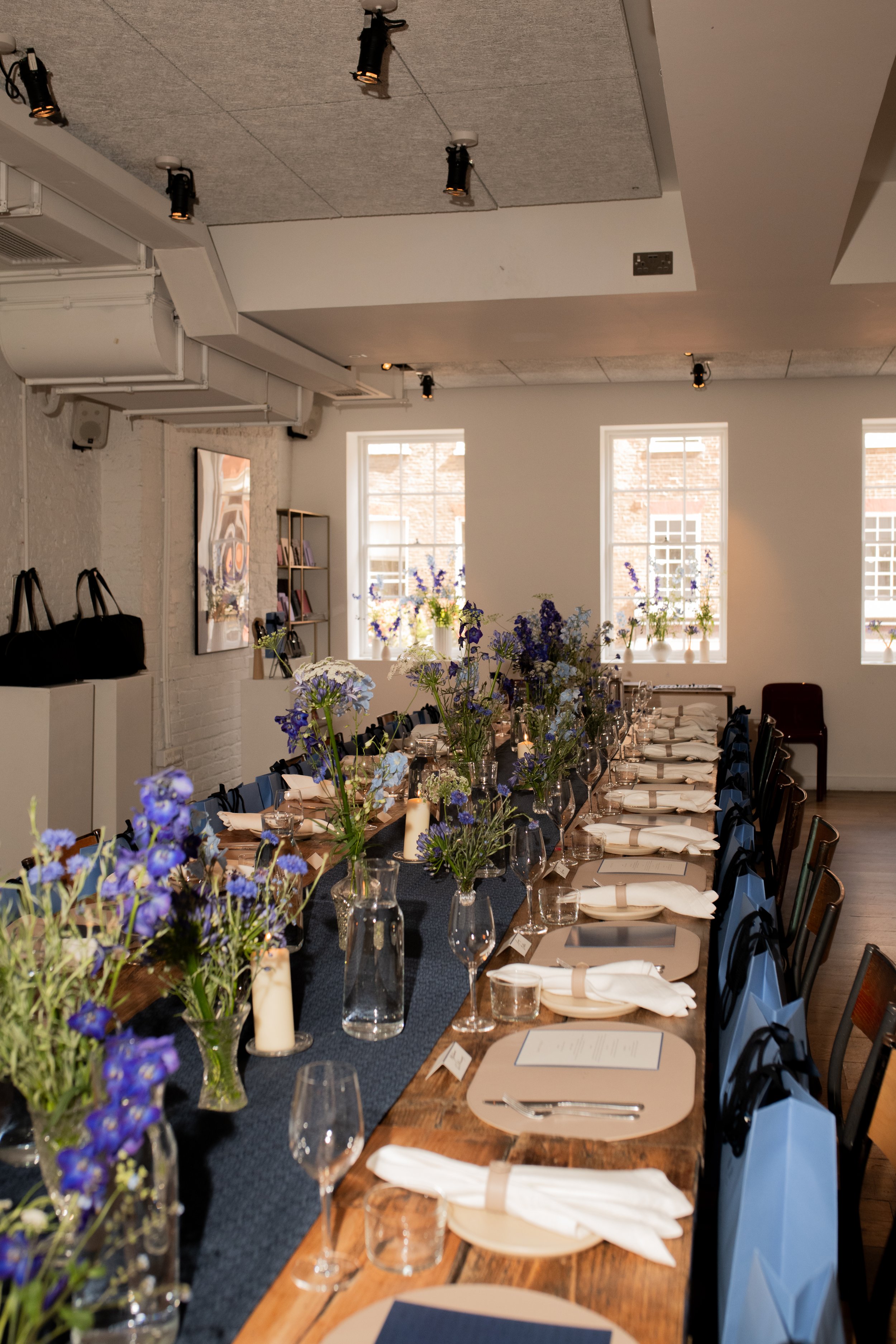A dining table set for a meal with floral centerpieces, candles, glassware, napkins, and place cards in a well-lit room with large windows and decorations.