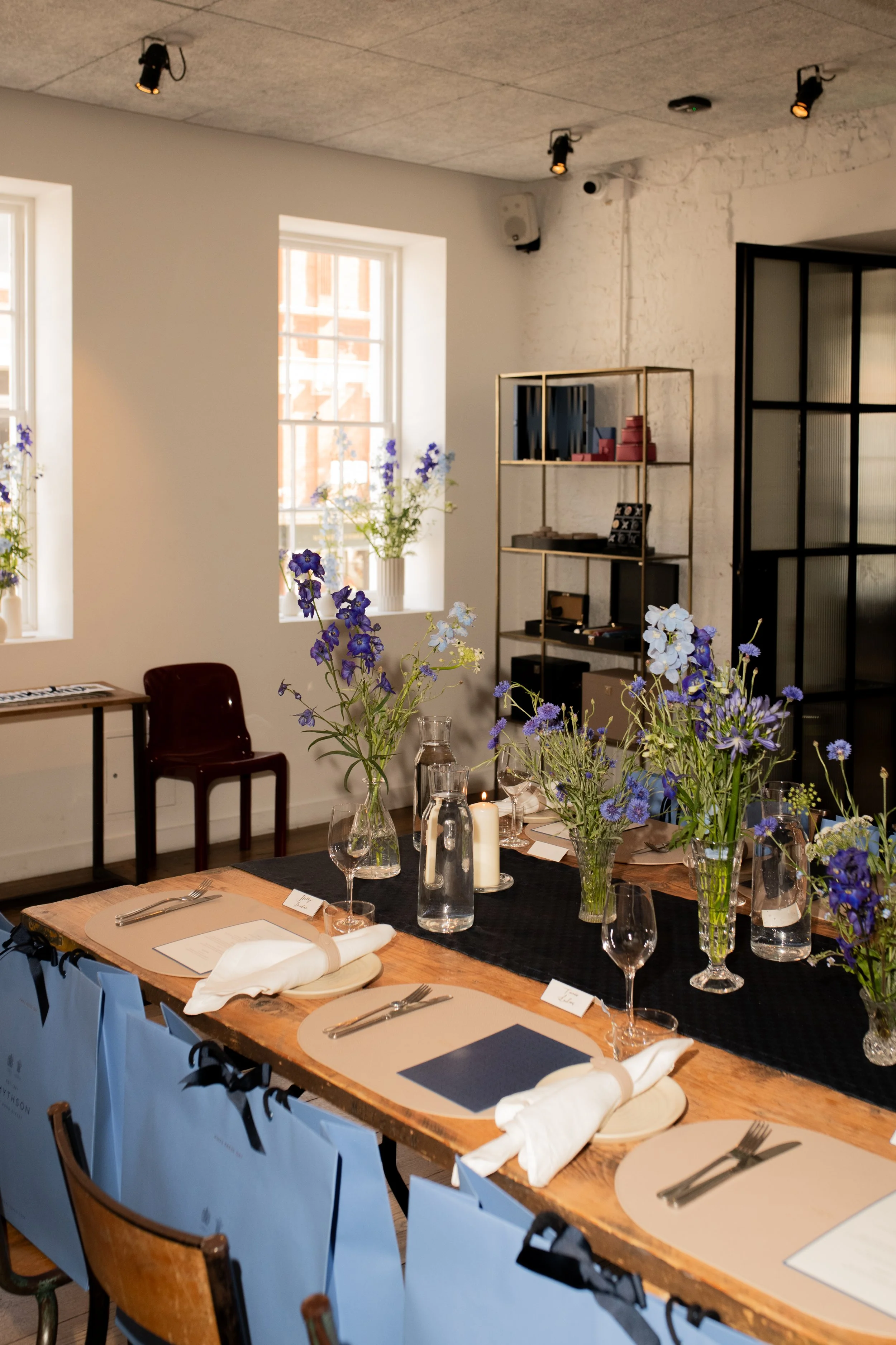 A dining table set for a meal with blue and white flowers in vases, candles, and place settings in a well-lit room with large windows, bookshelves, and decorative elements.