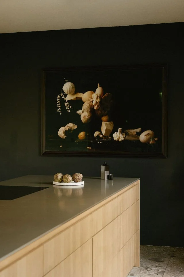 A modern kitchen with a light wooden countertop and a dark wall. On the countertop, there is a white plate with several round, brown baked goods. There is a bottle and a small container, possibly for salt or pepper. A large, dark framed painting or photograph hangs on the wall, depicting a still life composition of various vegetables and herbs.