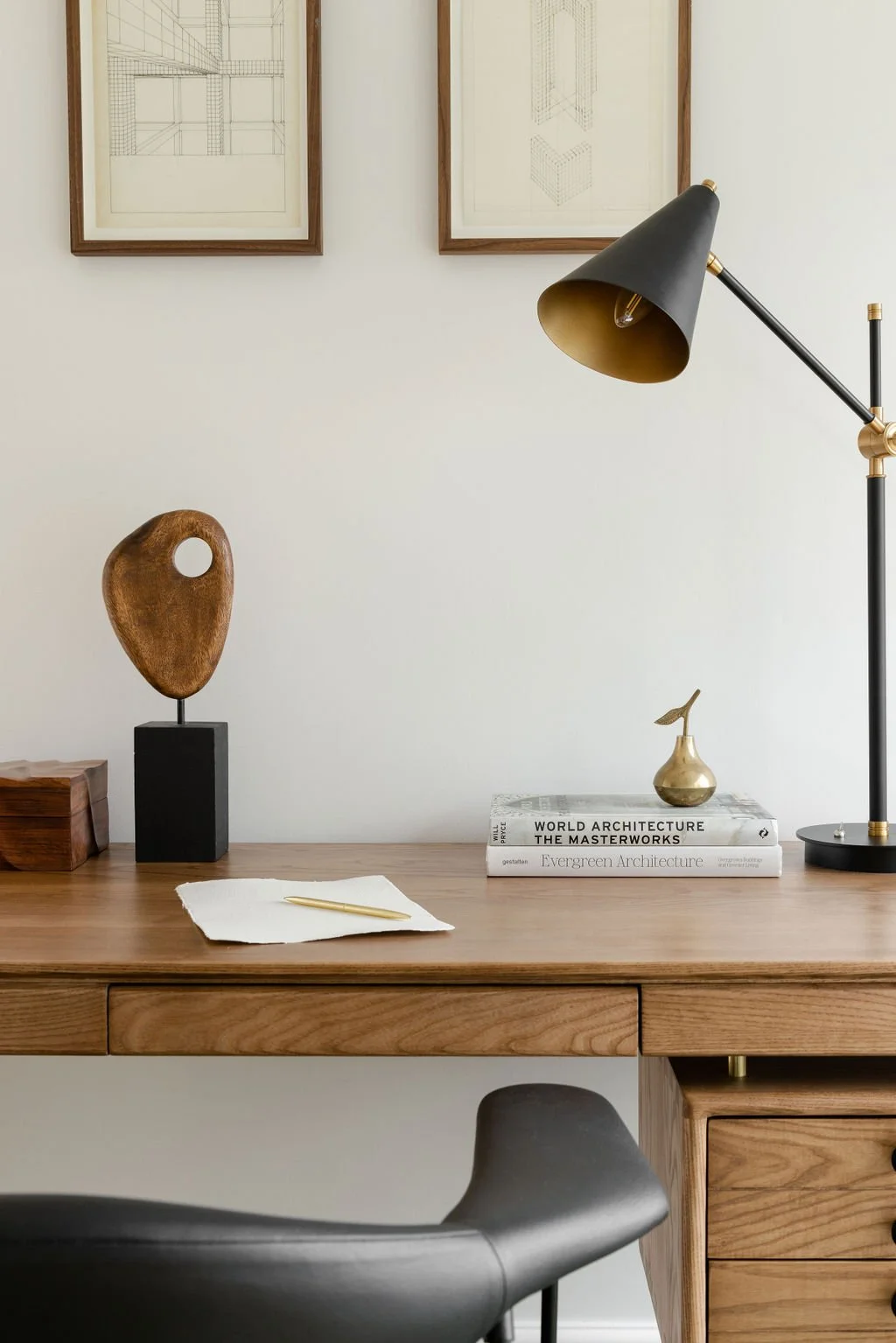 A modern wooden desk with decorative items, including a brass vase, a black and gold desk lamp, a small wooden sculpture on a black pedestal, a stack of architecture books, and a white cloth with a gold pen. Two framed sketches of buildings hang on a white wall above the desk.