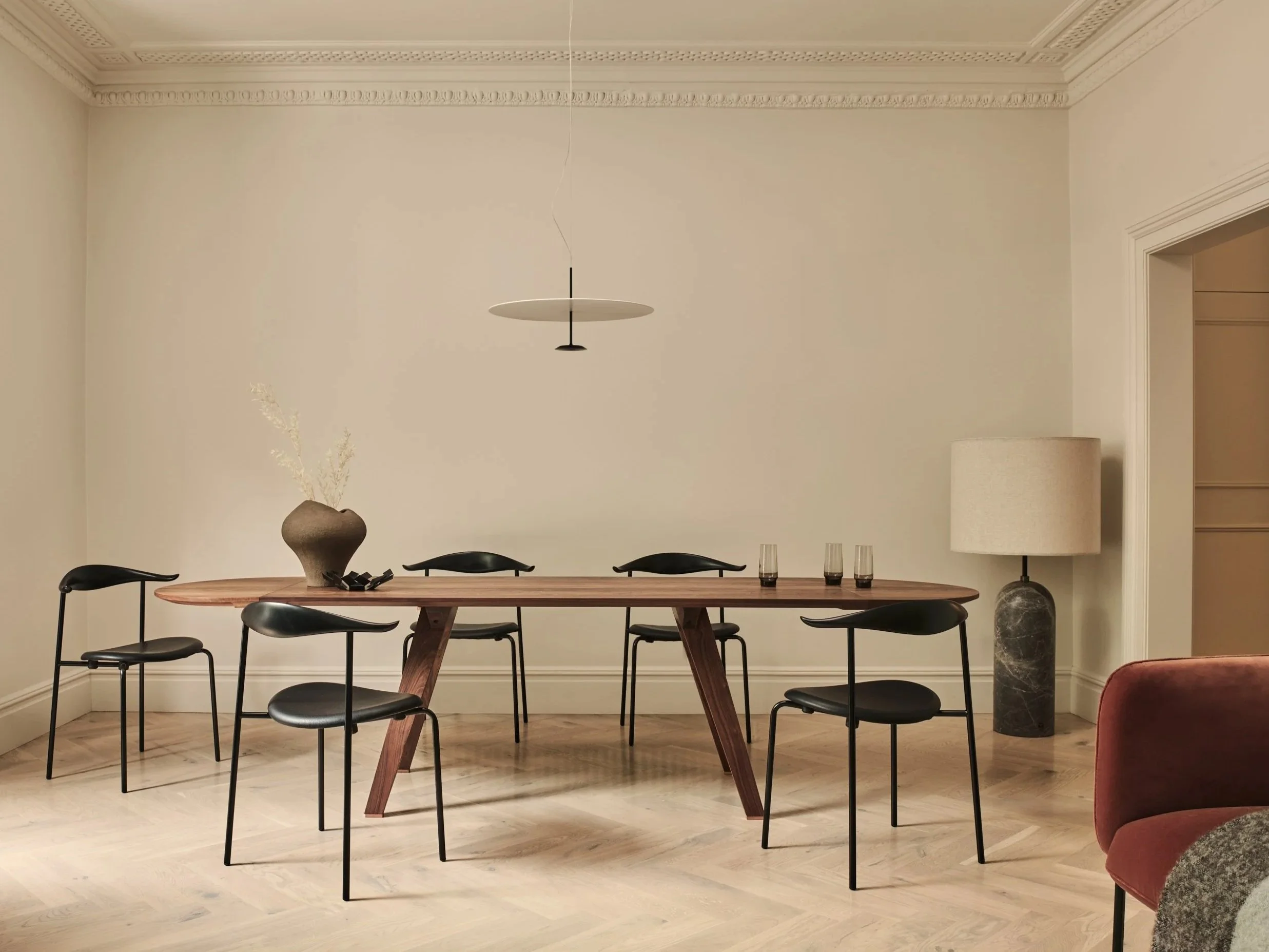 Modern dining room with a wooden table surrounded by six black chairs, a beige ceramic vase with dried plants, glasses on the table, a floor lamp with a beige shade and dark marble base, and a red upholstered chair partially visible in the corner.