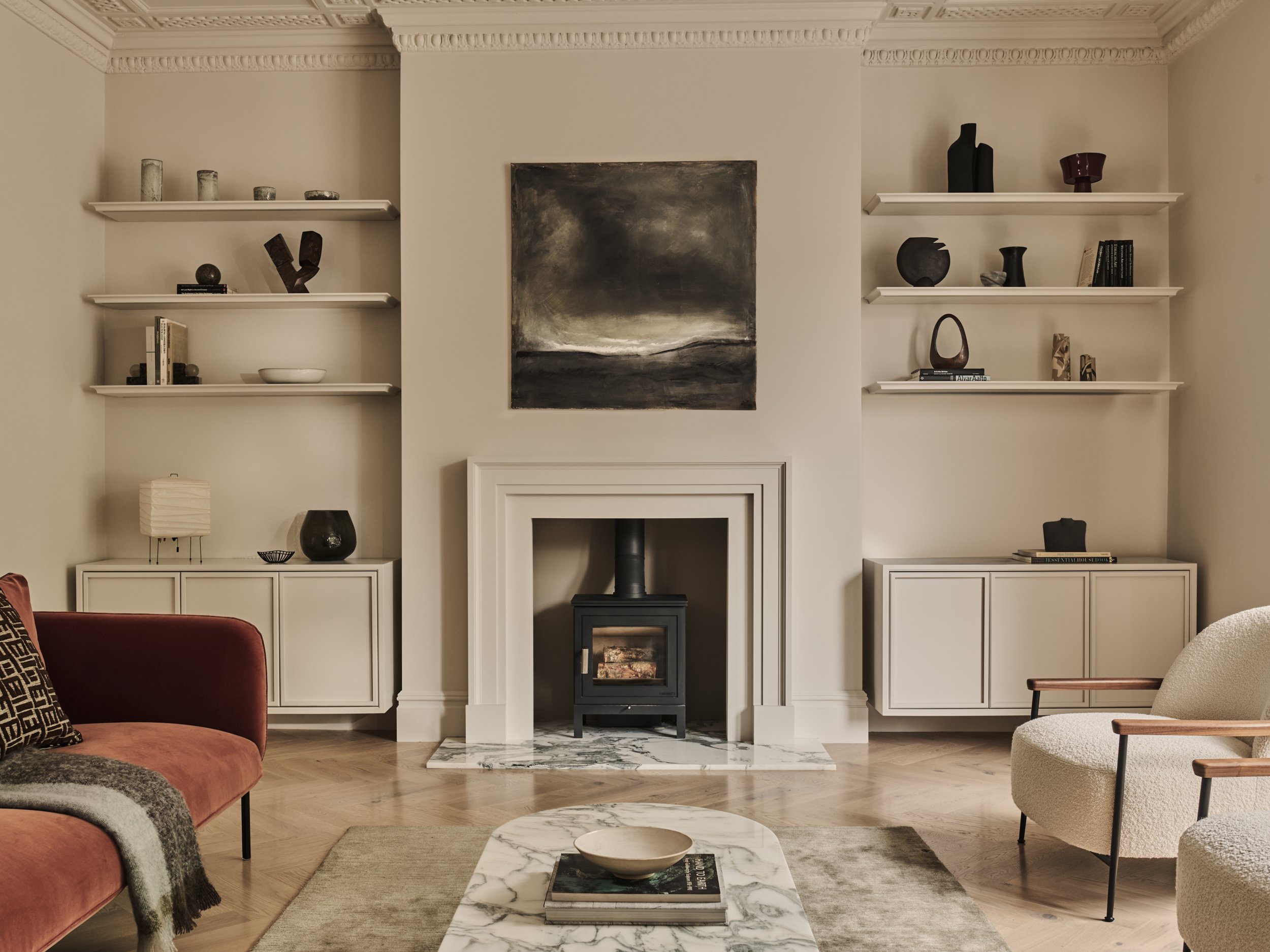 Living room with a fireplace flanked by shelves holding decorative objects and books, a painting above the mantel, and furniture including a pink armchair, white armchair, and a marble coffee table.