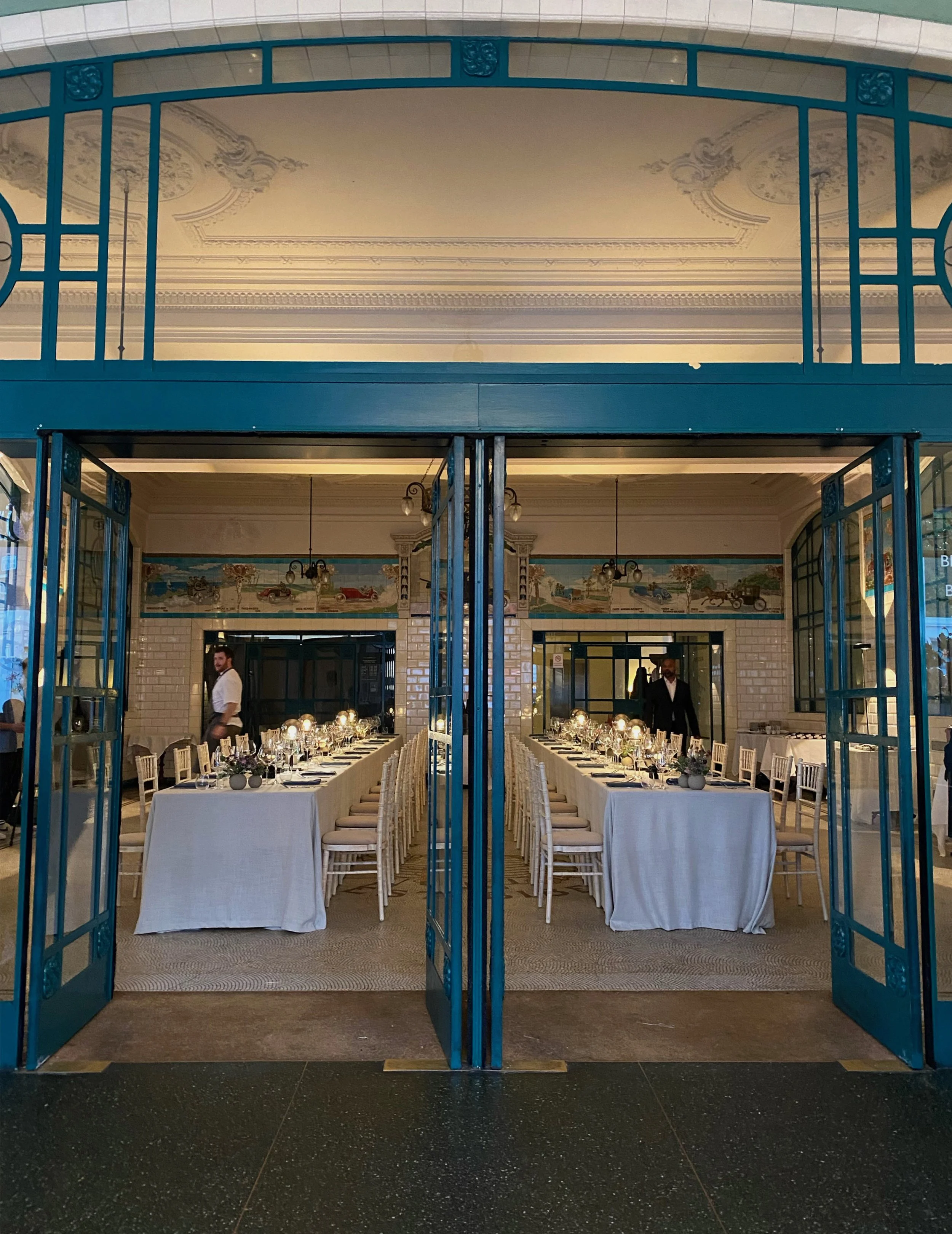 Elegant dining area set for a formal event with two long tables covered with white tablecloths, arranged with glassware, plates, and floral centerpieces. The room has high ceilings with decorative moldings, a mural on the back wall, and vintage-style