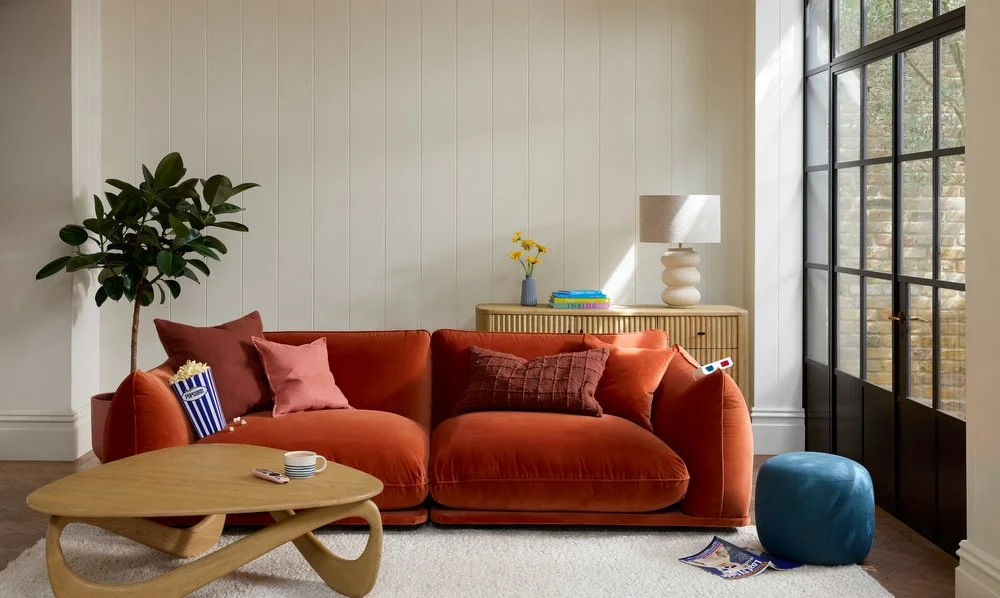 Living room with a red velvet sofa, a wooden coffee table, a tall potted plant, a striped popcorn box, a cup and remote on the table, a blue ottoman, and a wooden sideboard with a vase of yellow flowers, a lamp, and books. Large glass doors are on the right, letting in natural light.