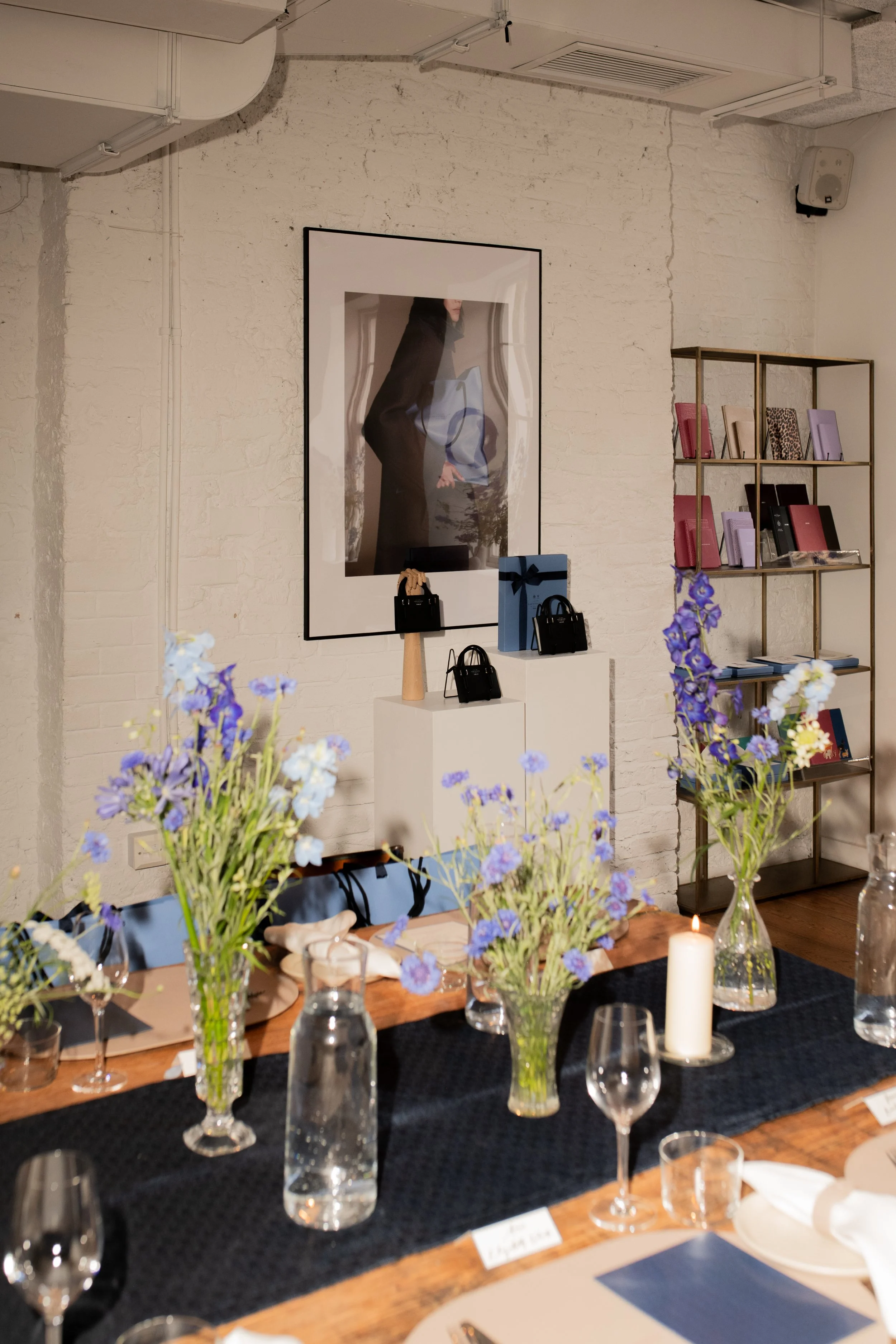 A decorated dining table with purple and white flowers in vases, a lit candle, wine glasses, water bottles, and place settings. In the background, shelves hold colorful folders, and a large framed artwork hangs on a white brick wall.