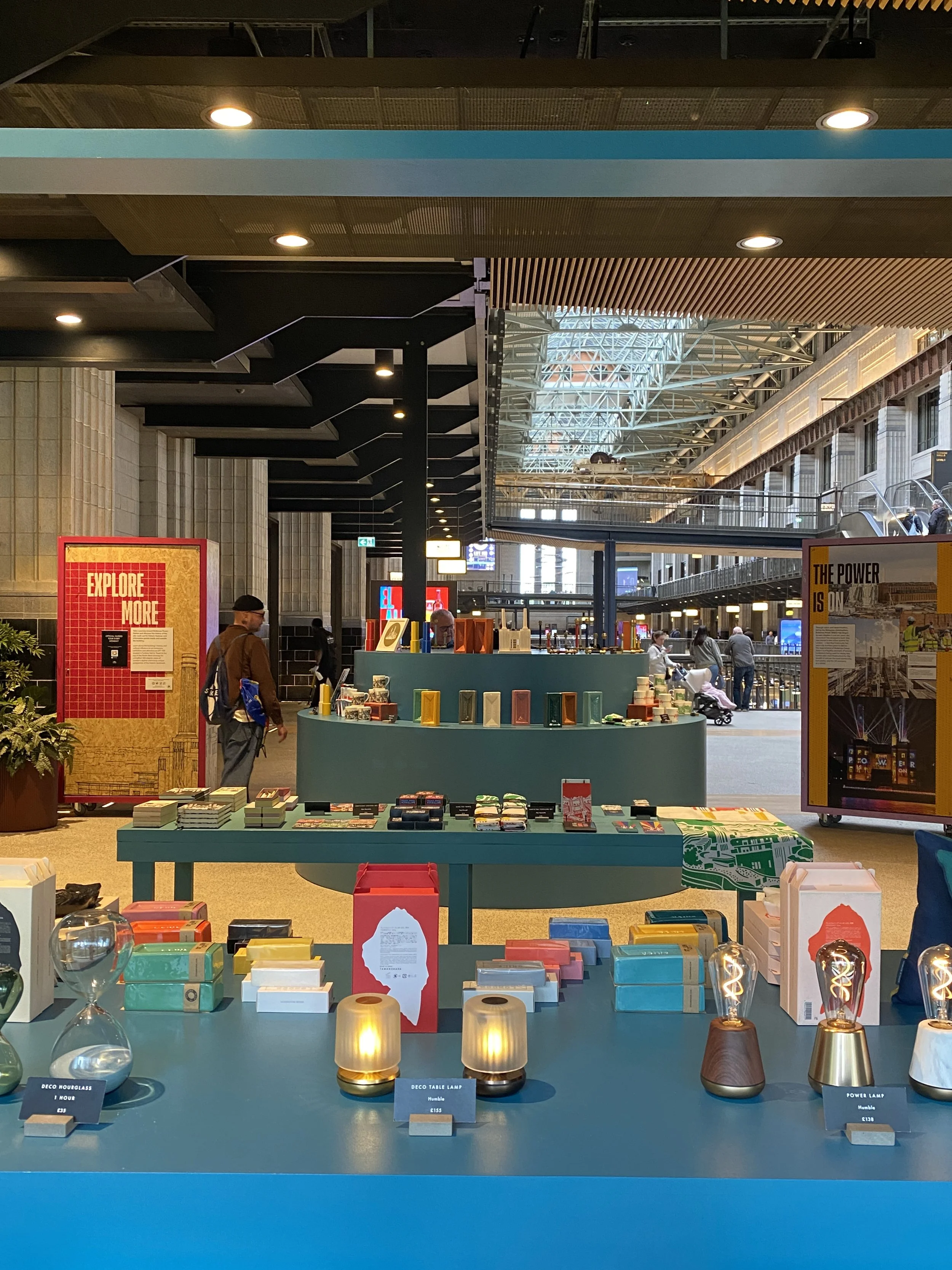 Display of various modern lamps and home decor items on blue tables at a retail or exhibition space in an industrial-style building.