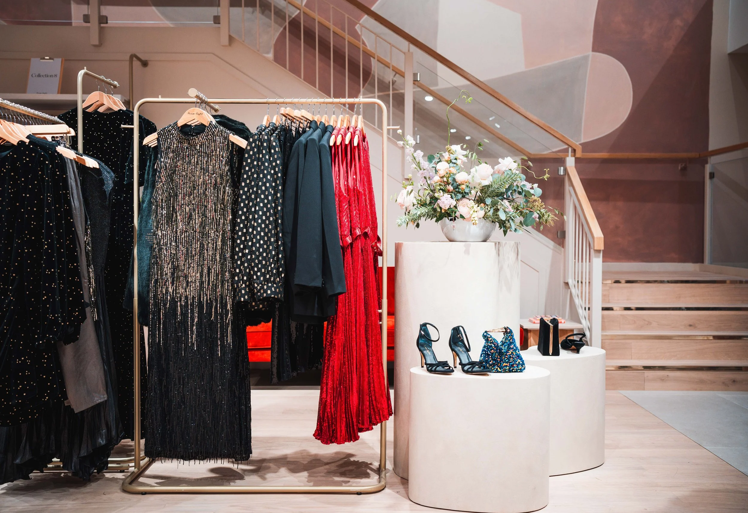 Clothing rack with elegant black and red dresses, and jackets, next to a display with black high heels, a patterned bag, and a black clutch, all on round white pedestals, and a large floral arrangement in a white vase in a modern boutique store.