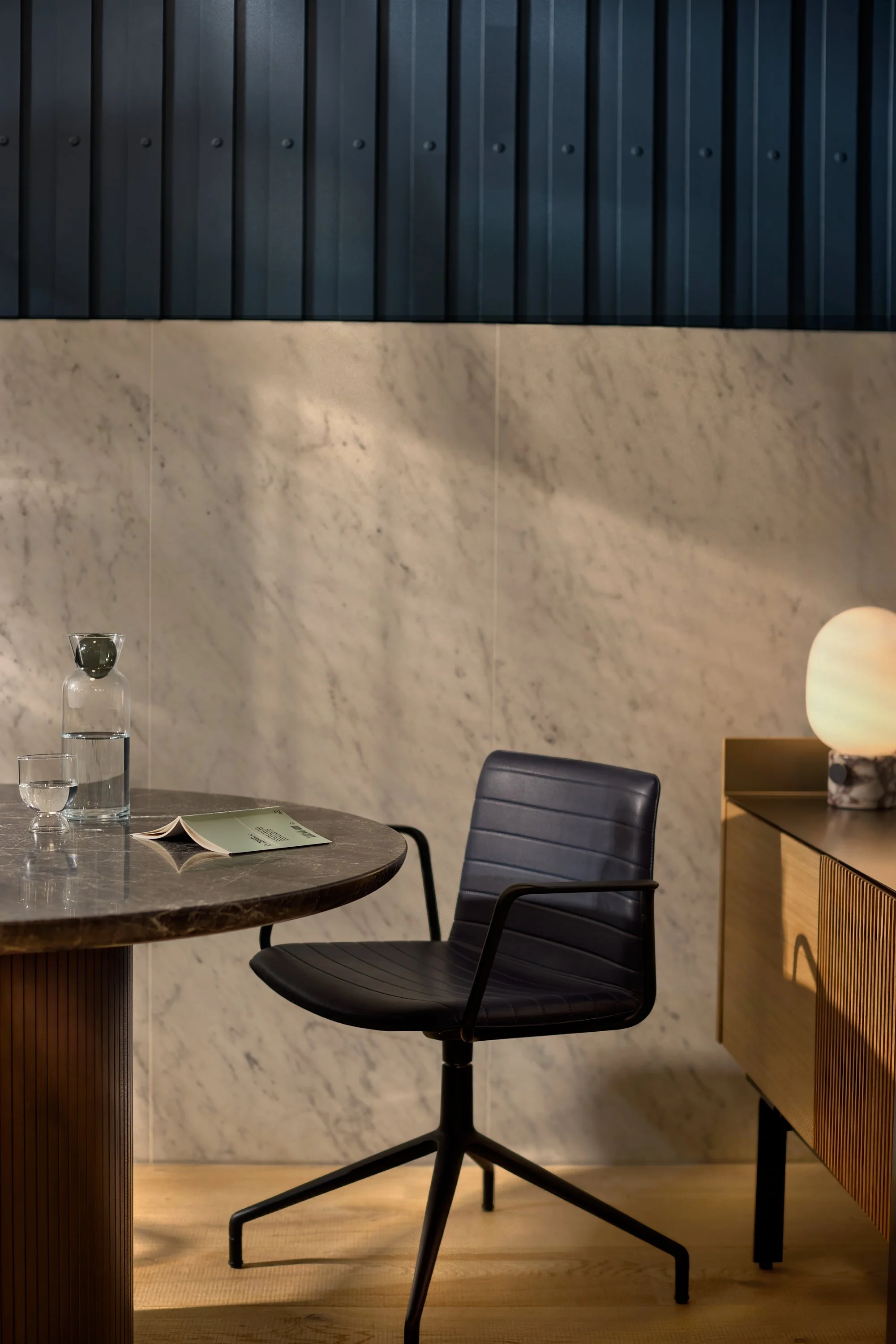 A modern office or meeting room with a black office chair, a marble table with a glass water pitcher, a glass of water, and an open book. There is a wooden sideboard with a lamp and a large marble base, and a dark wall with vertical panels in the background.
