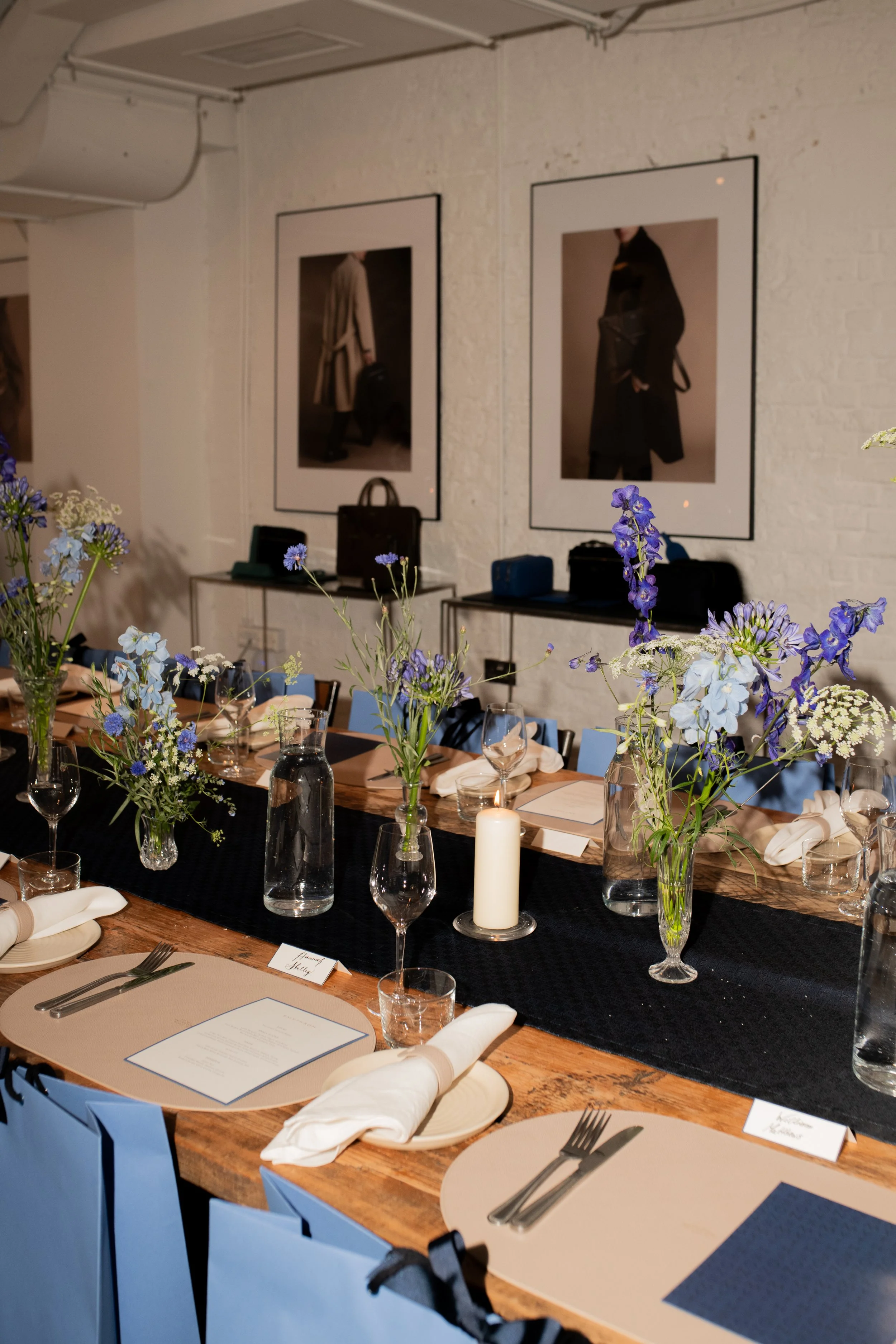 Decorated dining table with blue flowers in vases, candles, wine glasses, plates, and silverware, set for a formal event in a room with artwork on the white brick wall.