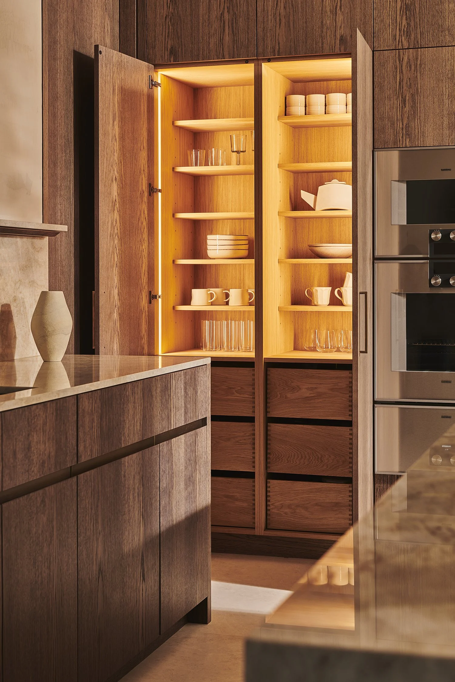 Open wooden cabinet with illuminated interior, displaying stacked dishes, glasses, mugs, and bowls in a modern kitchen.