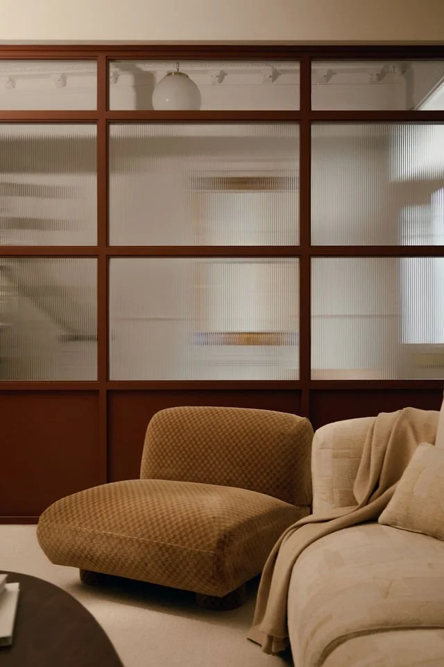 A beige armchair and a beige sofa with a throw blanket in a living room, with a wooden-framed glass partition wall behind them.