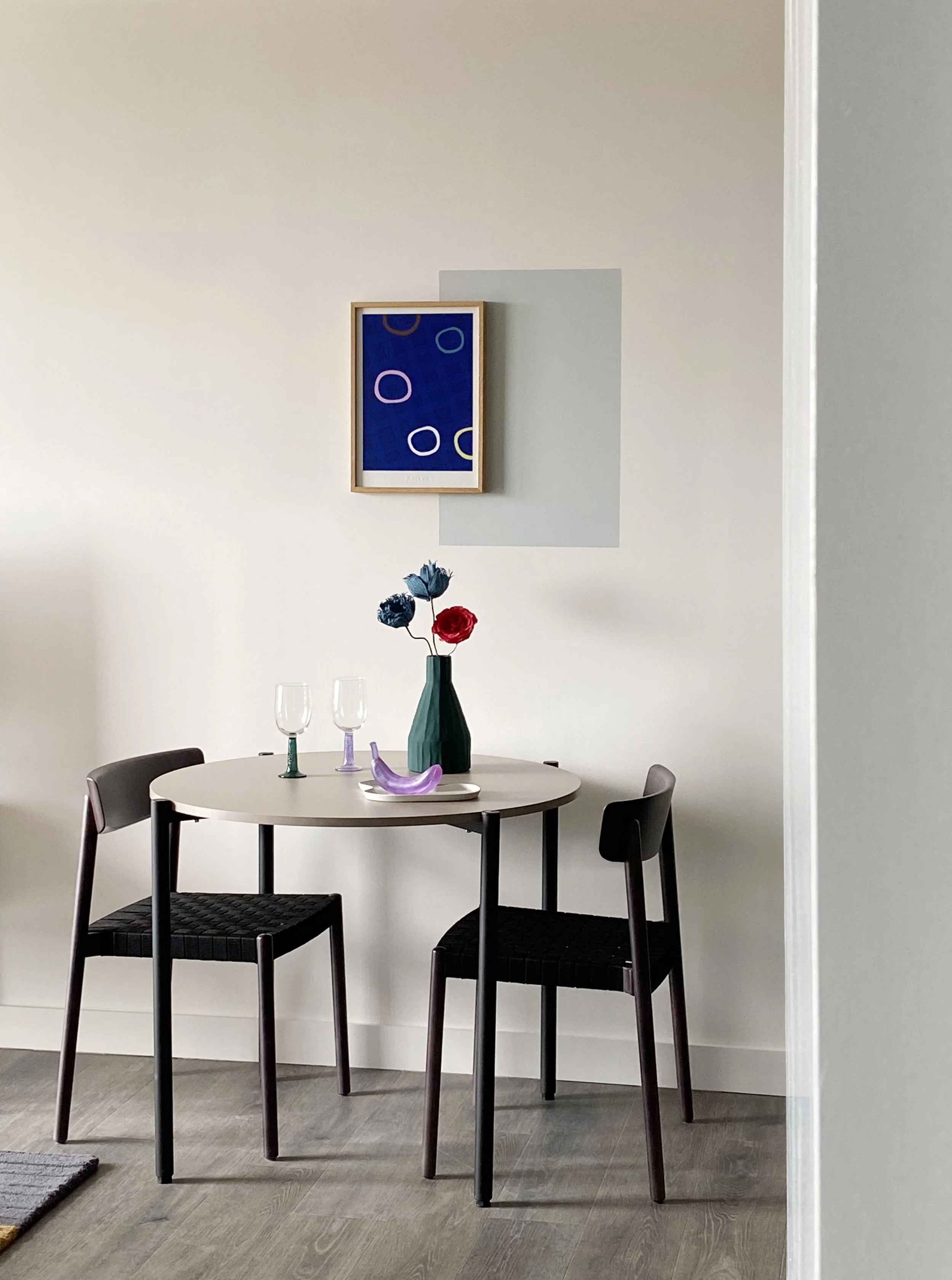 A small round dining table with four black chairs, a green vase with blue and red flowers, two wine glasses, and a purple decorative object, placed against a white wall with a colorful abstract painting.