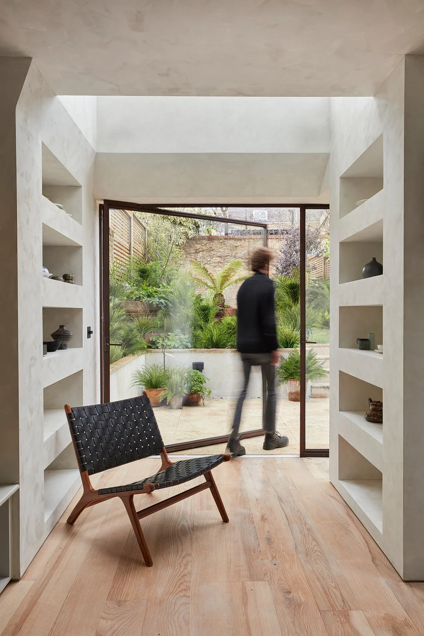 Interior view of a room with built-in white shelves on either side, a wooden floor, and a large sliding glass door opening to a lush garden with various green plants. A blurred person is walking outside the door.