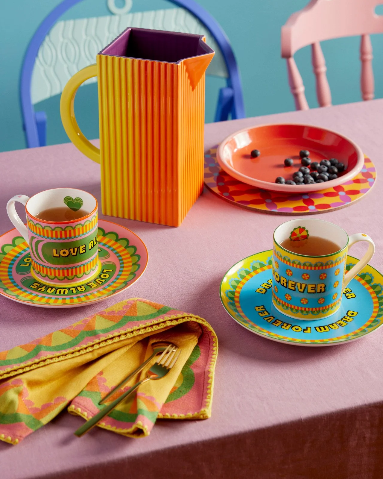 Colorful table setting with two cups of tea on patterned saucers, a pink bowl with blueberries, a large orange mug with a purple interior, a multicolored napkin with a fork and spoon, all placed on a pink tablecloth with pastel chairs in the backgrou