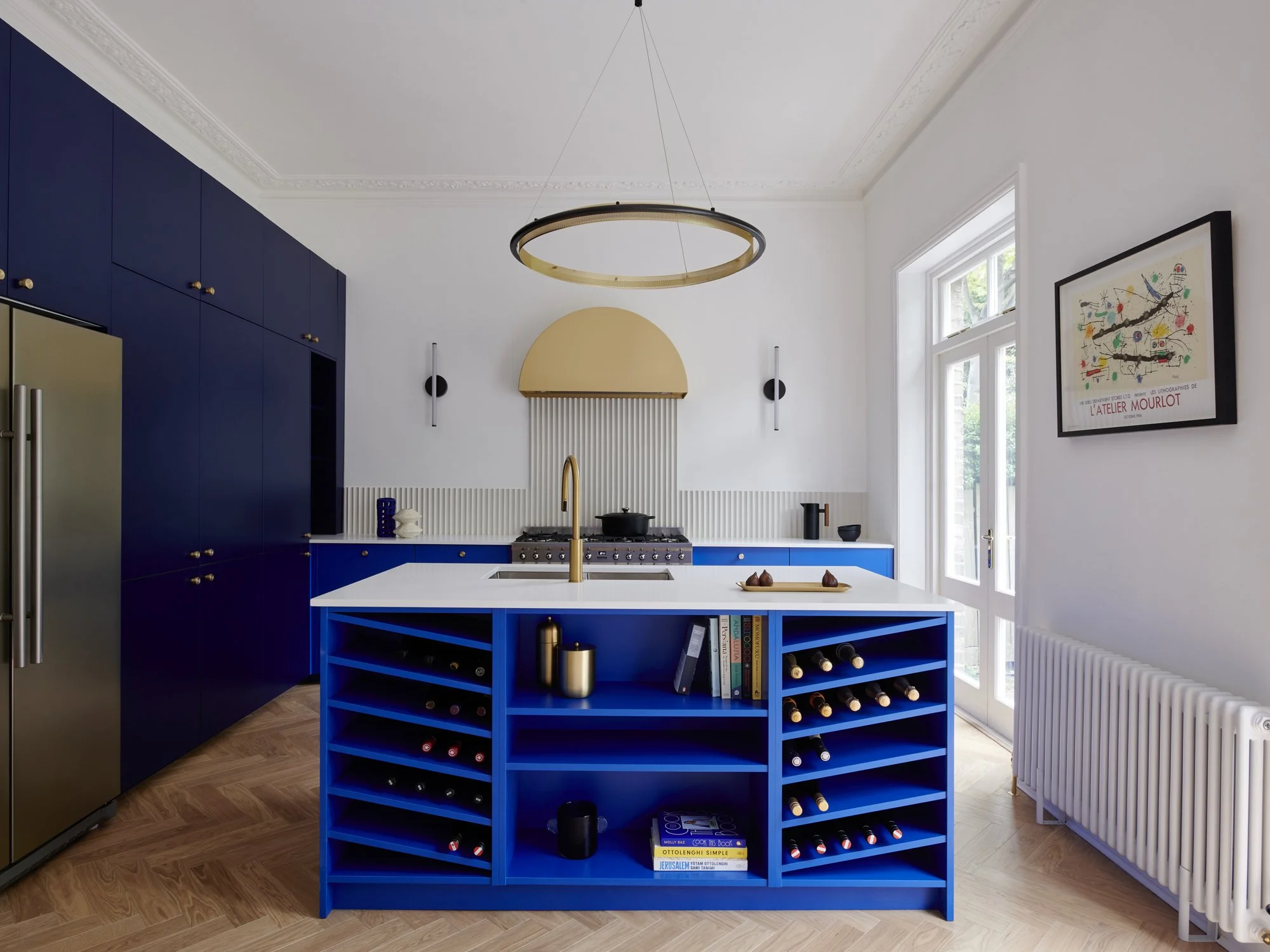 Modern kitchen with navy blue cabinets, a blue wine rack island, white countertops, and a gold faucet. Includes a window, framed artwork, and a gold modern ceiling light.