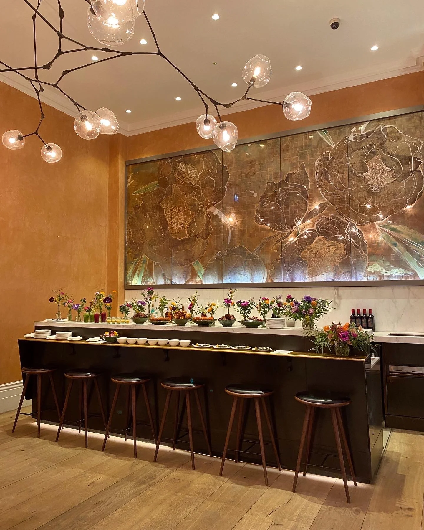 A decorated buffet table with flowers, bowls, and bottles, set against a brown and gold floral wall in a dining area with wooden chairs and a modern chandelier.