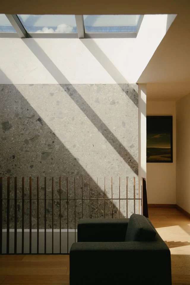 Interior view of a modern building with a large, angled window casting shadows on a textured stone wall and wooden floor, with a green armchair and a wooden railing in the foreground.