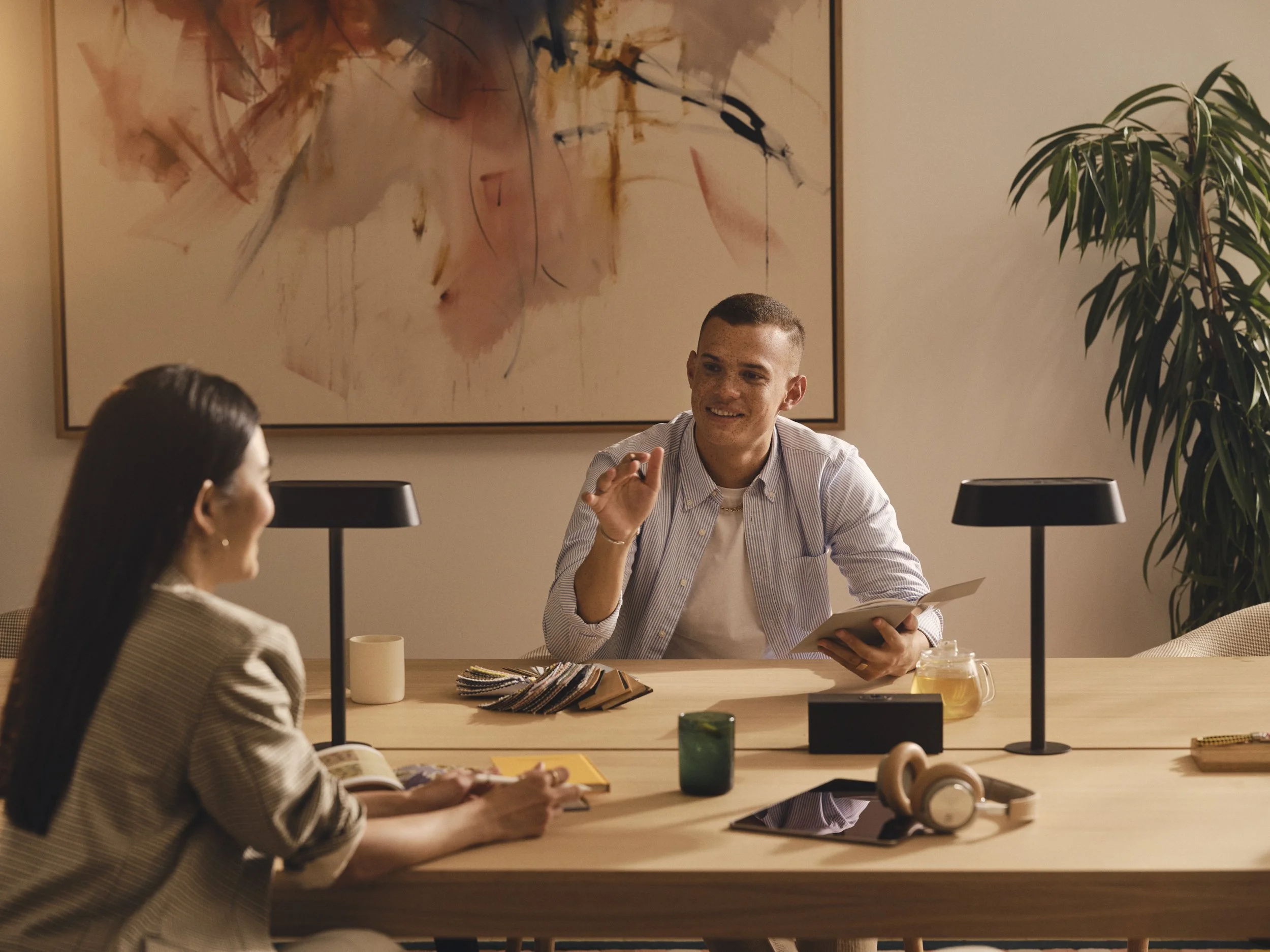Man and woman having a conversation at a work meeting in a modern office, with abstract art on the wall, a large plant, and various office items on the table.