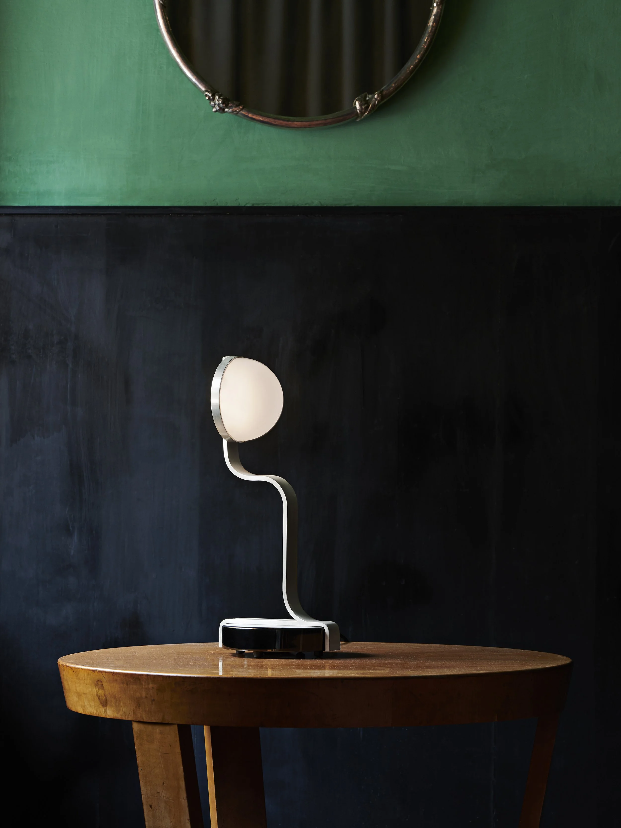 Table lamp with a curved stand and round shade on a wooden table against a dark wall with a green upper section and mirror above.