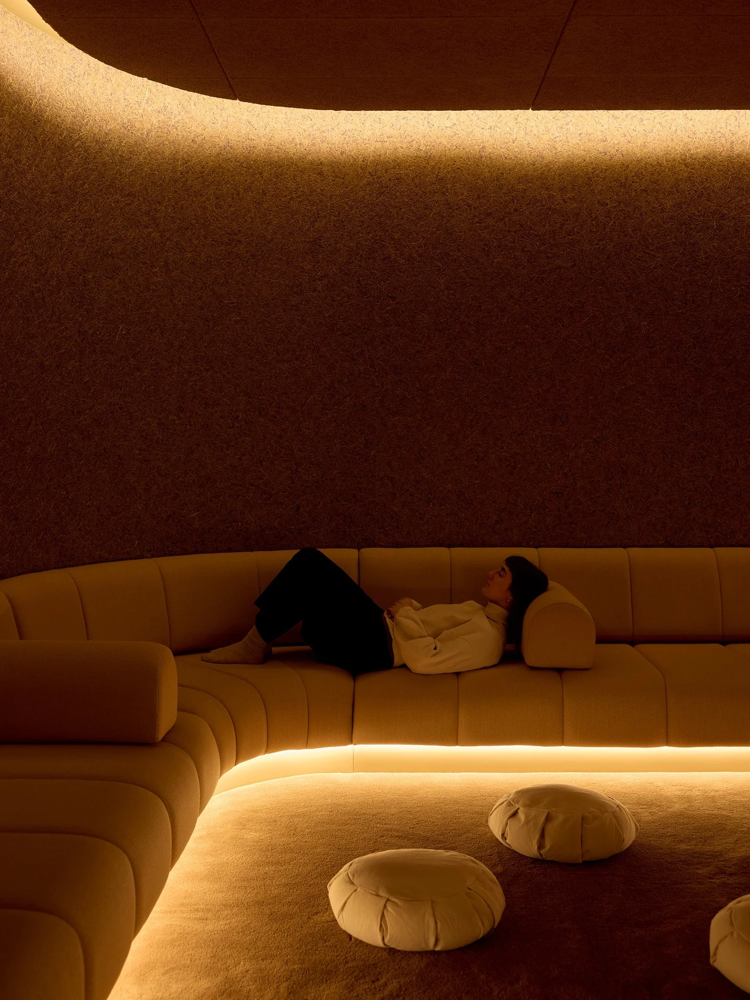 A woman lying on a curved beige sofa with a built-in headrest, in a dimly lit room with a textured brown wall and soft, warm lighting, and four beige poufs on the floor.