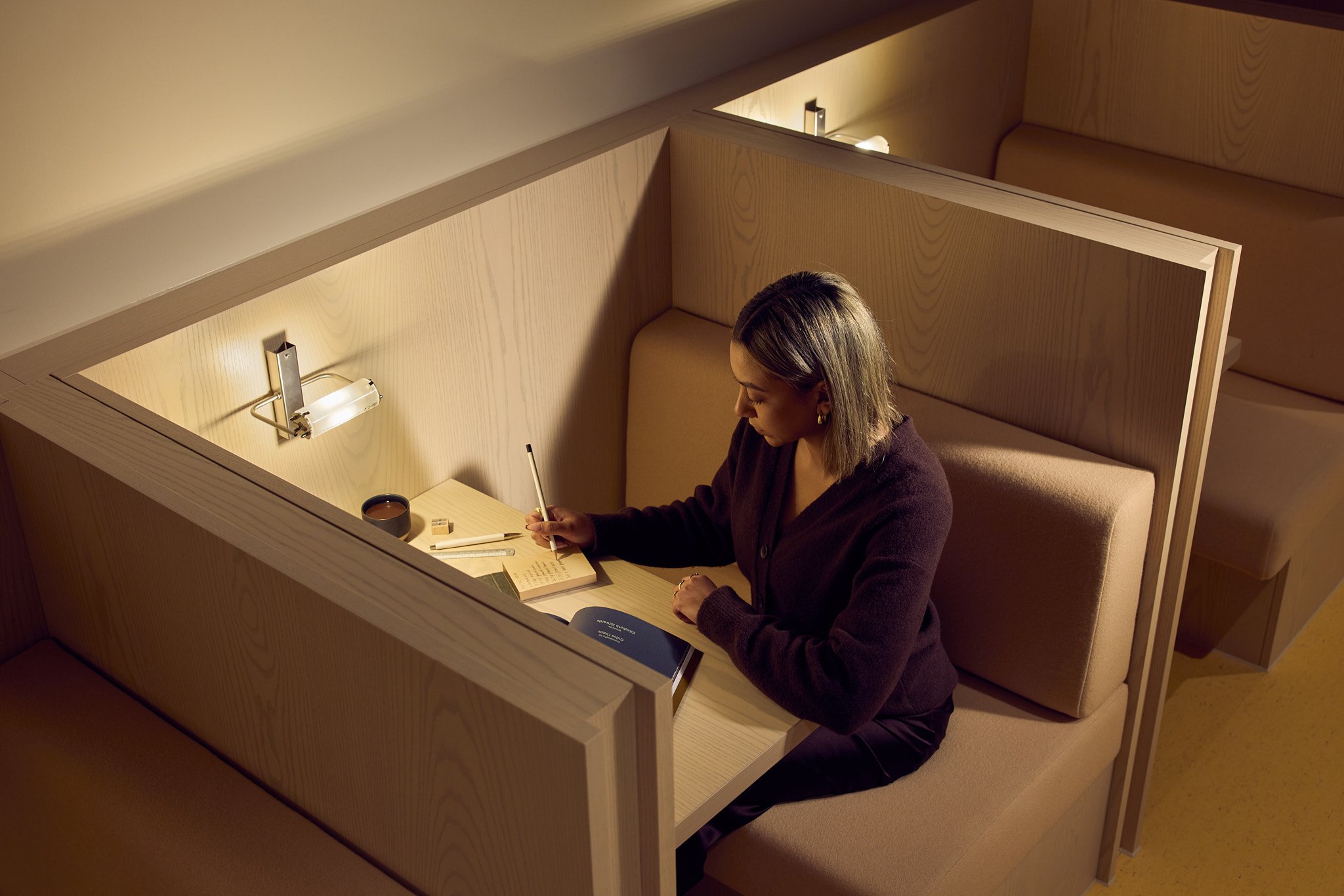 A woman sitting in a private wooden booth, writing in a notebook with a cup of coffee, surrounded by study materials and a book, illuminated by wall-mounted lighting.