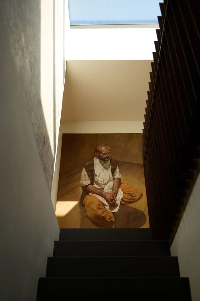 Inside a modern building, looking upward at a landing with a large photograph of an elderly man in traditional clothing, sitting cross-legged on the floor, with natural light from a skylight above.