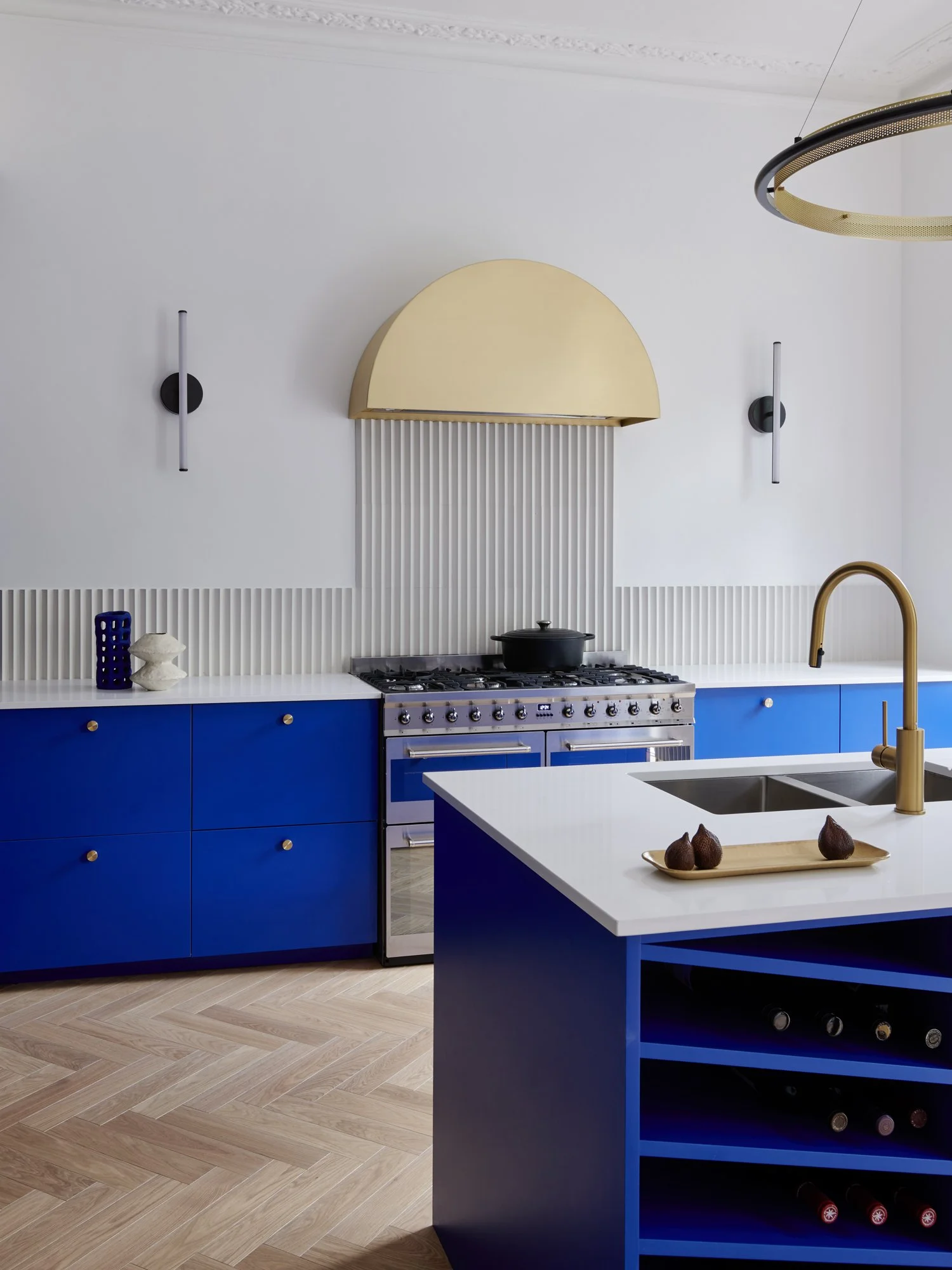 Modern kitchen with blue cabinets, white countertops, and a gold faucet. There is a dish with three brown objects on it, a black stove with a pot, and decorative items on the counter. A large gold semicircular vent is above the stove, and black and w