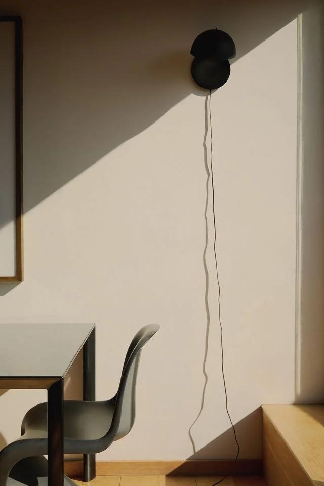 Black wall lamp with a long cord casting a shadow on a white wall in a room with a table and chair.