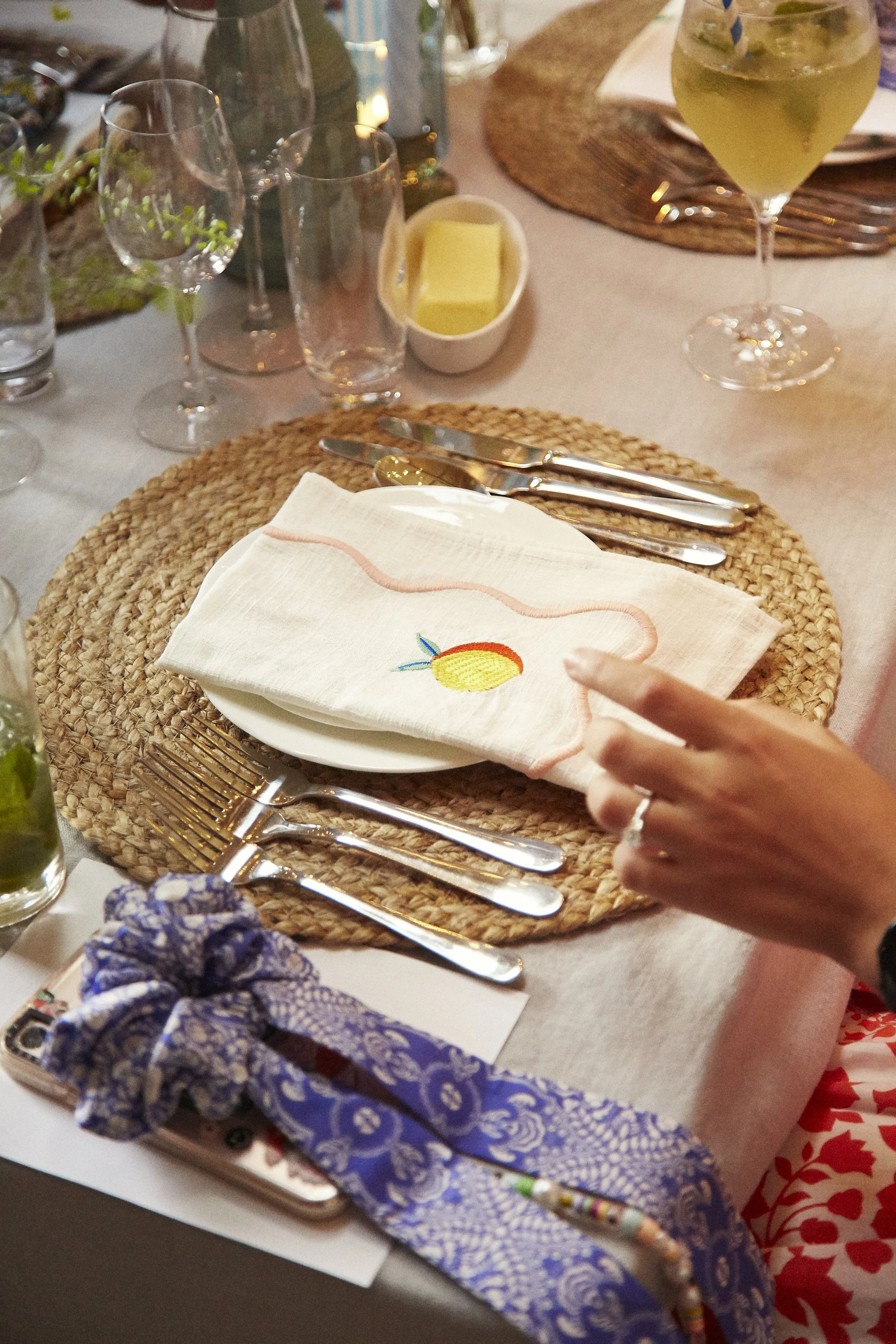 A table setting with a woven placemat, white plate, embroidered napkin, silverware, and a hand with a ring. There are glasses and a drink in the background, along with a butter dish and a decorative cloth on a phone.