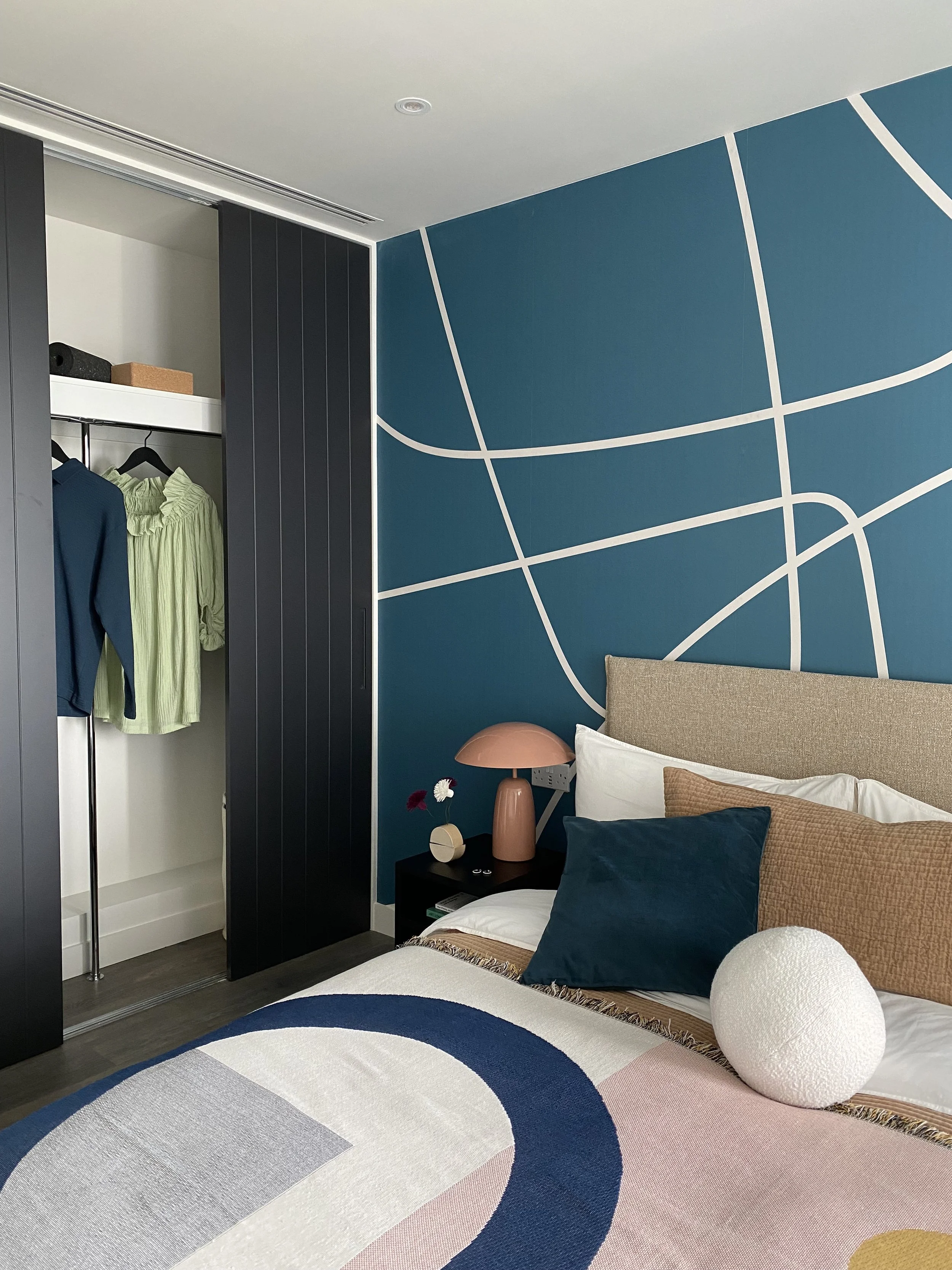 A bedroom with a blue wall featuring white curved lines, a beige headboard, a black nightstand with a pink lamp, and a bed with pillows and a blanket. An open closet with hanging clothes, including a green blouse and a blue jacket, is visible on the 