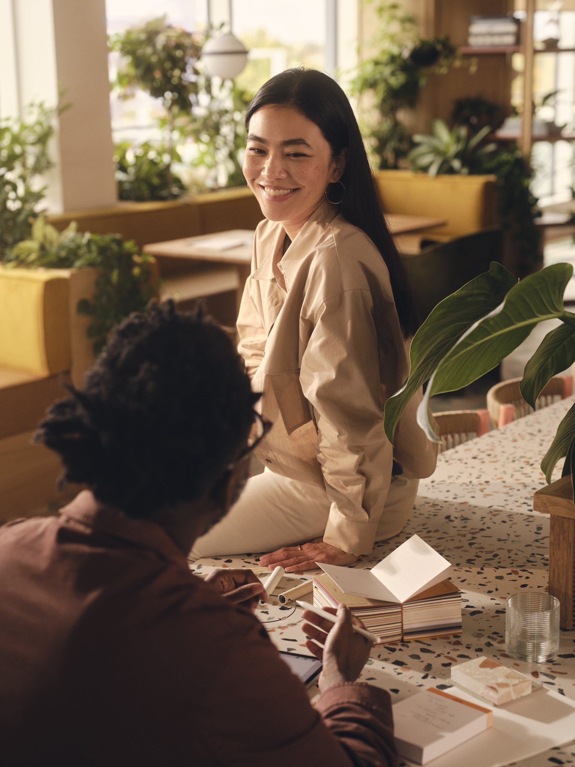 A woman with long dark hair smiling while sitting on a table, engaging in conversation with a person with curly dark hair and glasses in a cozy, plant-filled room with warm lighting.