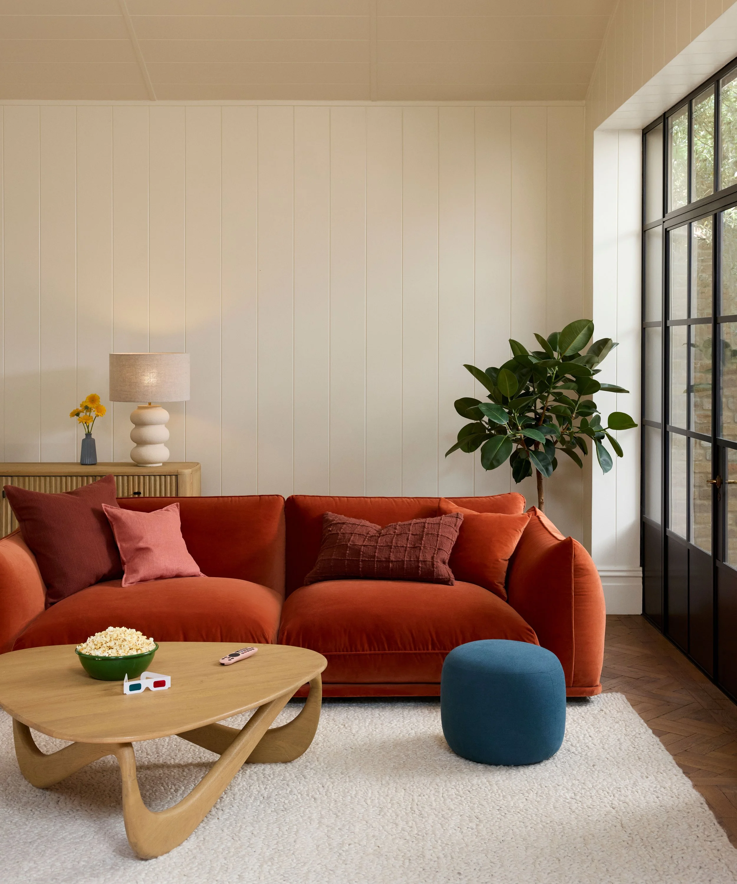 Living room with a red sofa, wooden coffee table with popcorn, a blue ottoman, a potted plant, large window, and a side table with a lamp and yellow flowers.