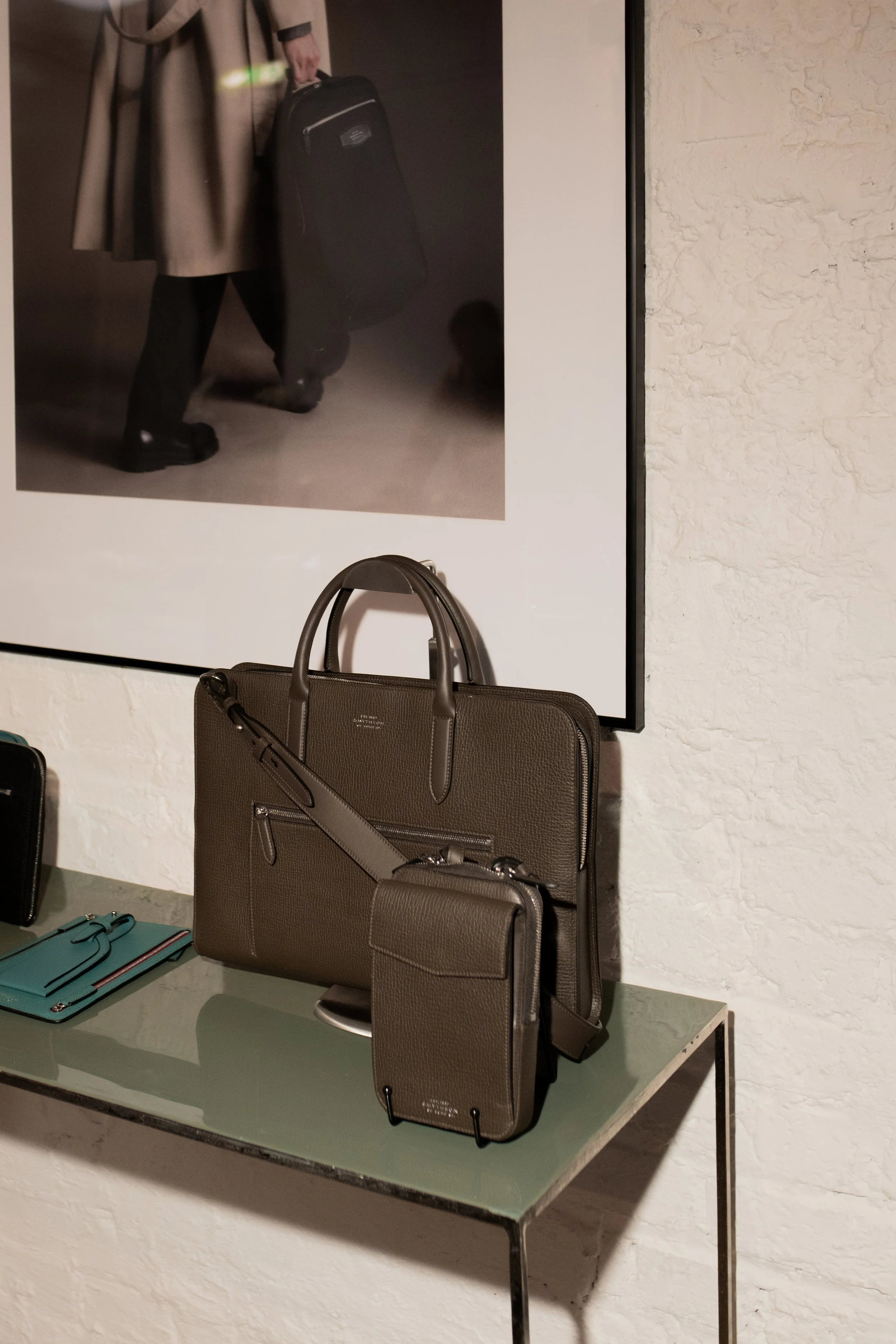 Display of designer handbags and accessories on a glass table against a white textured wall, with a framed black-and-white photograph on the wall behind.