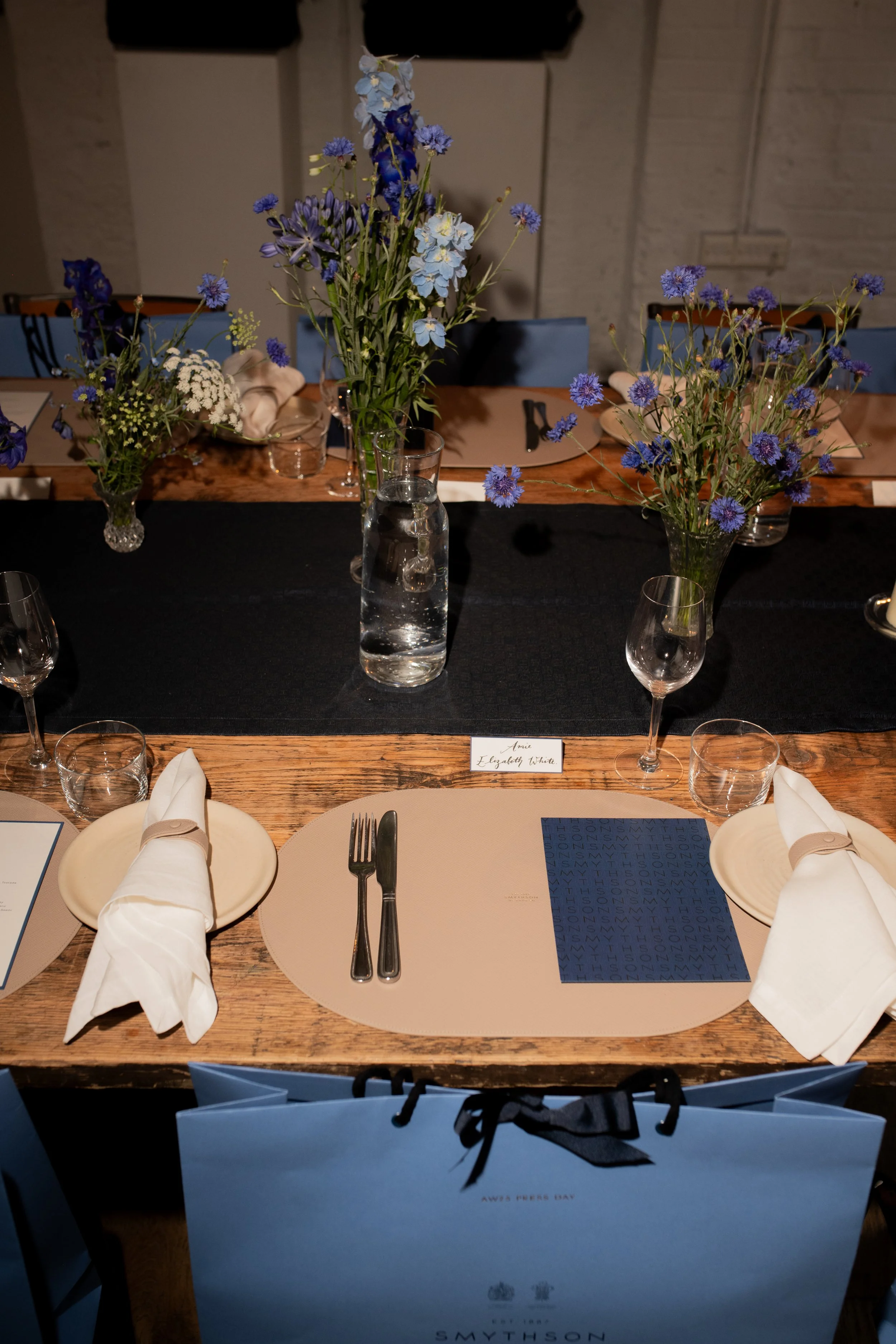 Table setup for a formal event with floral centerpieces, placemats, napkins, utensils, glasses, and branded gift bags. The flowers are primarily blue and white. A name card reads 'Annie Elizabeth White'.