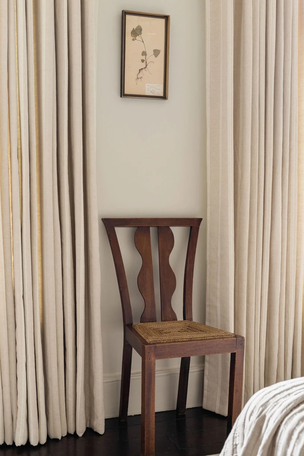 Empty wooden chair with woven seat between cream-colored curtains and a framed botanical print on the wall.