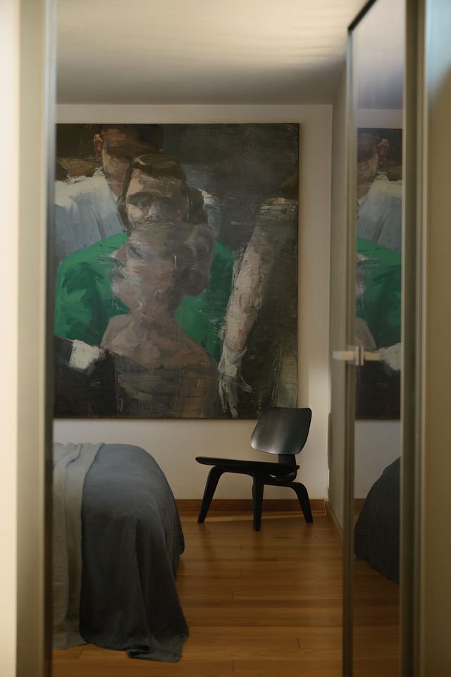 View of a bedroom with a large abstract painting of three people on the wall, a black chair, and part of a bed with gray sheets, seen through a mirror reflection.