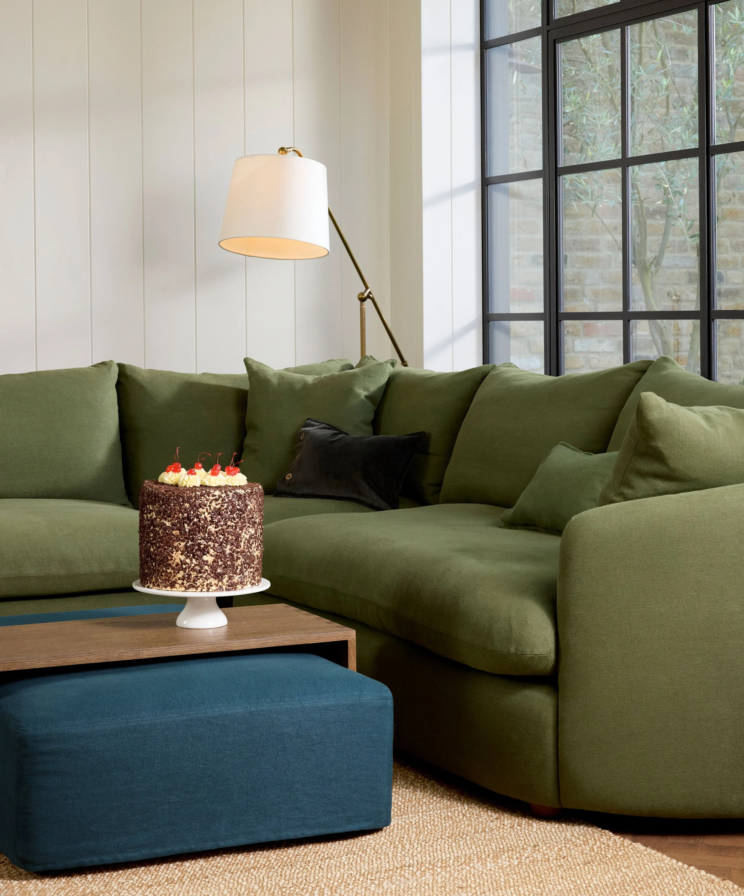 A living room with a green sectional sofa and black pillows, a wooden coffee table with a chocolate birthday cake topped with cherries and whipped cream, a lamp, and a large window.