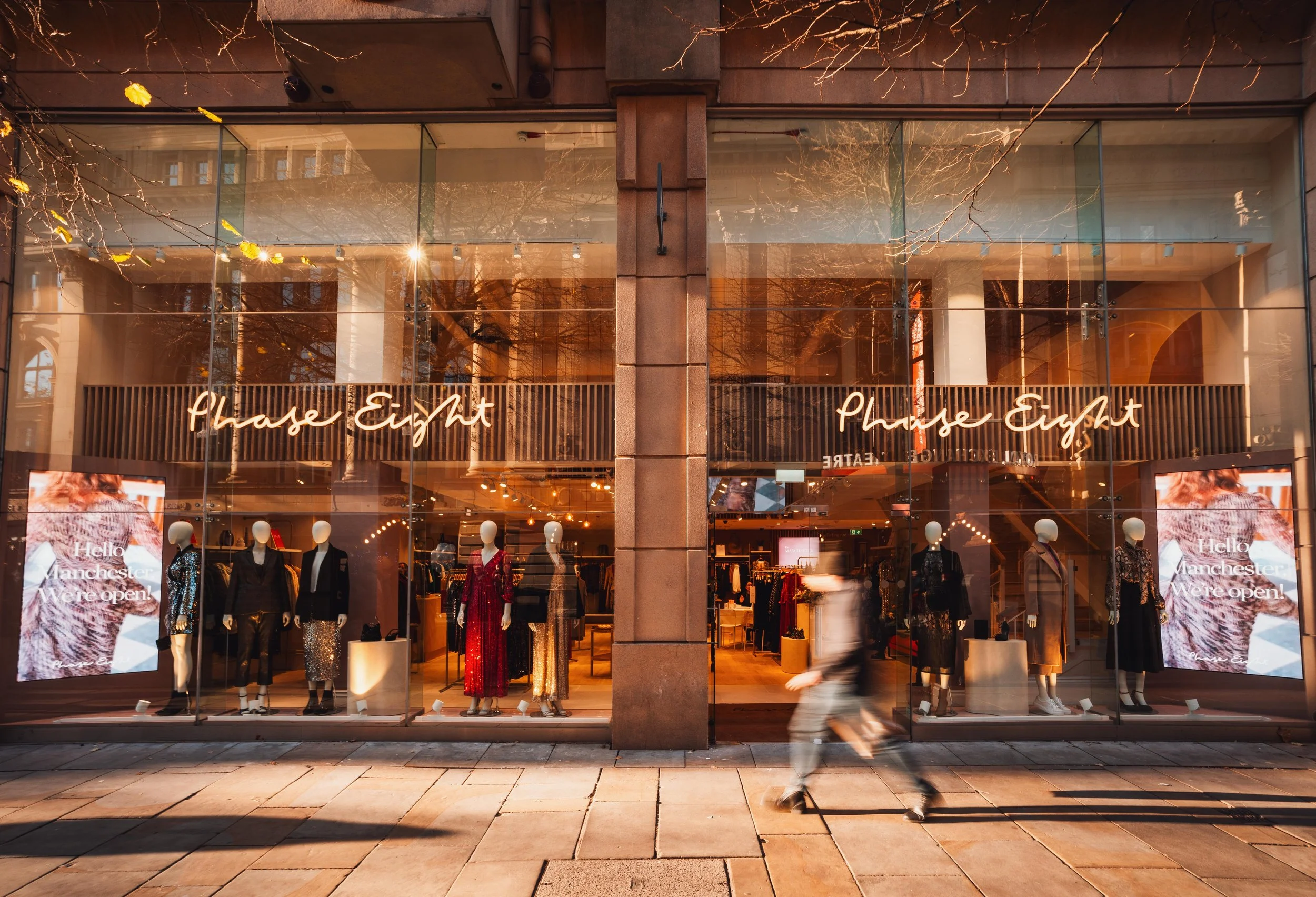 A storefront window display for a clothing store named 'Phase Eight'. Mannequins dressed in various outfits are displayed inside. A person is walking past on the sidewalk, and digital screens on either side of the window show a message about the stor