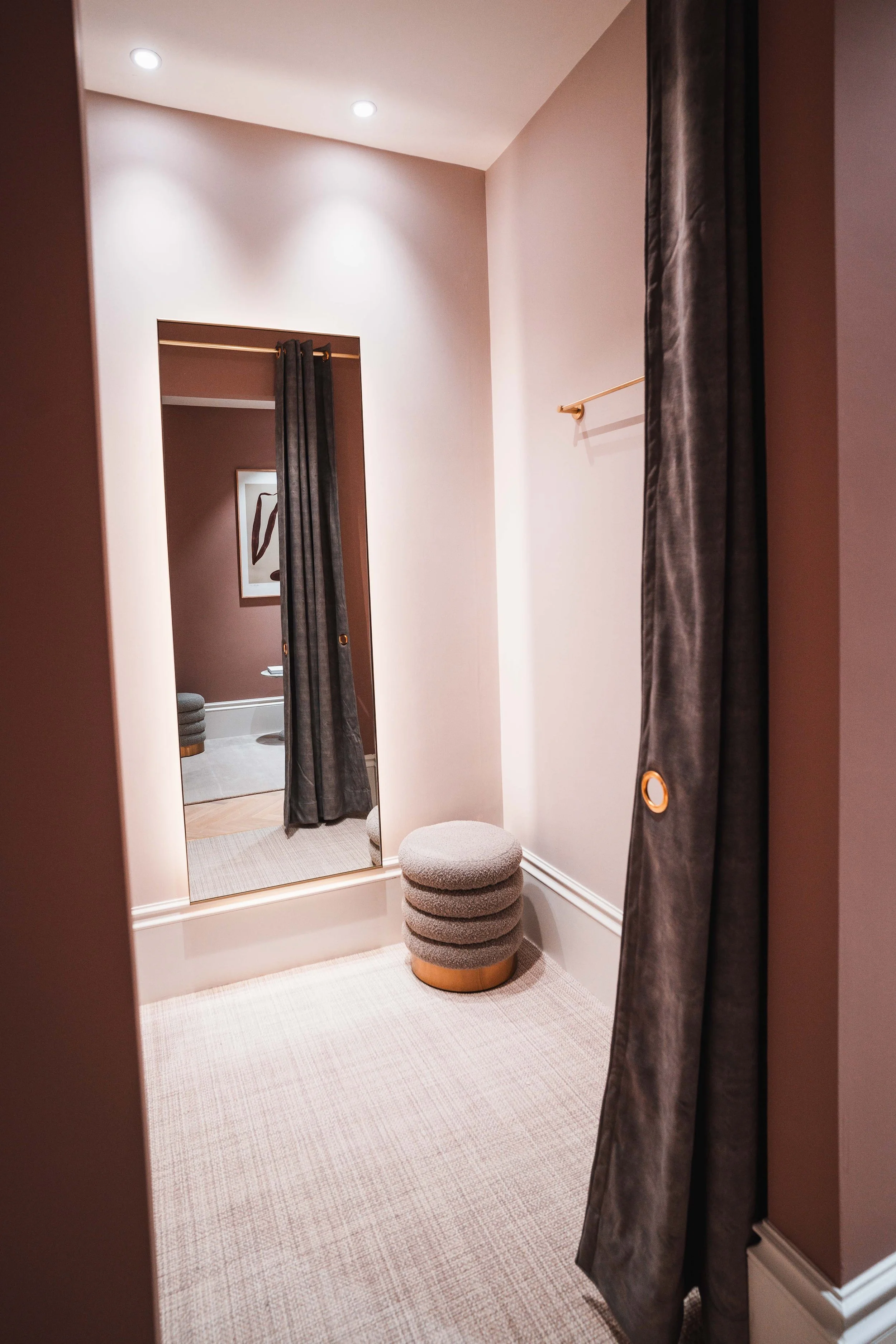 A fitting room with a full-length mirror, a small gray upholstered stool, and dark curtains, with soft lighting and a framed artwork on the adjacent wall.