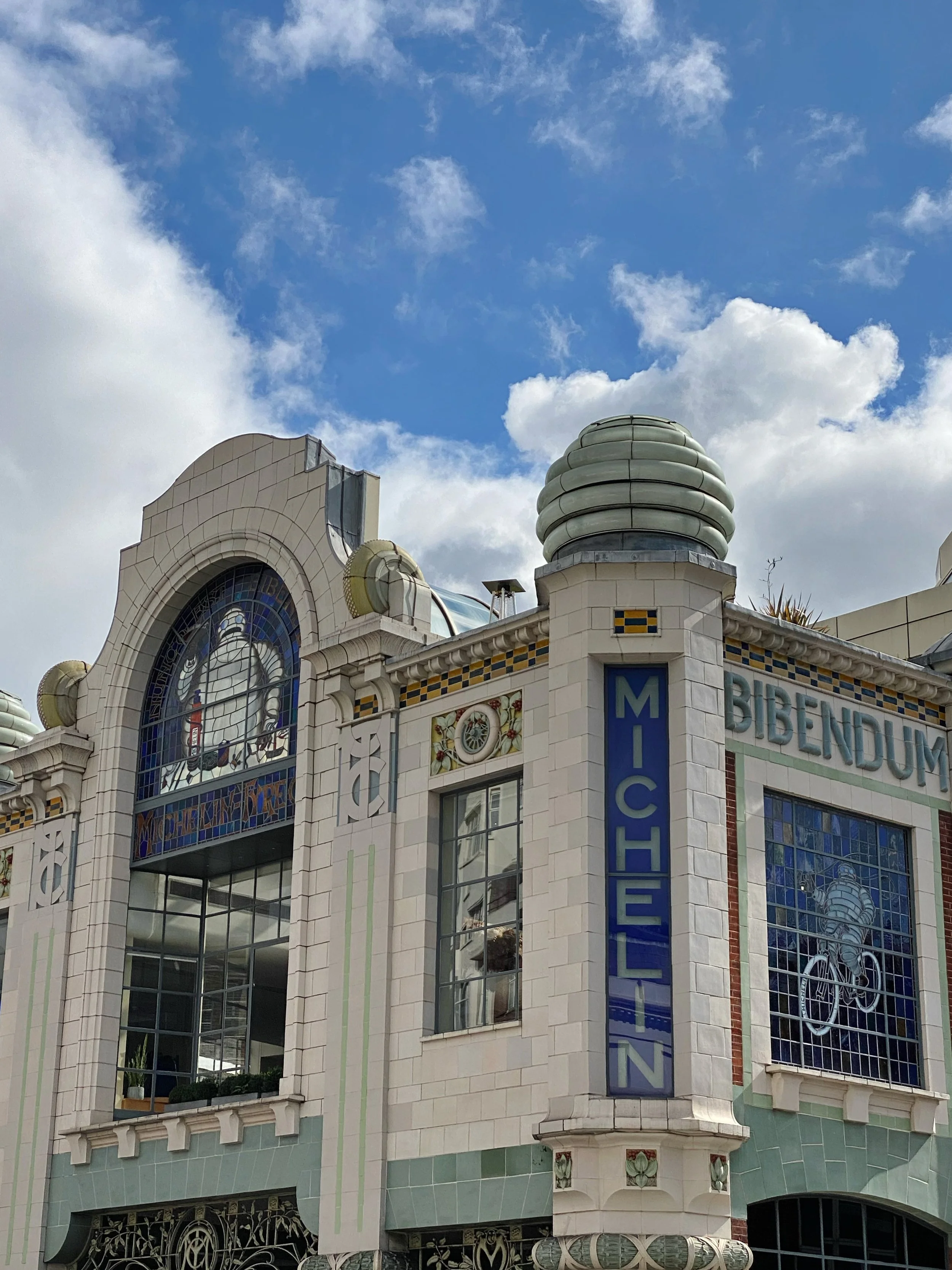 A building with decorative stained glass windows, a sign that reads 'MICHEL' and 'BIBENDUM,' and architectural features like rounded glass domes, under a partly cloudy blue sky.