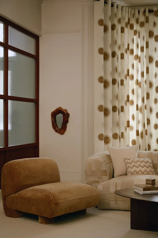 A living room corner with a beige patterned armchair, a light-colored sofa with throw pillows, a dark wooden coffee table with books, a decorative wall mirror, and floral patterned curtains.