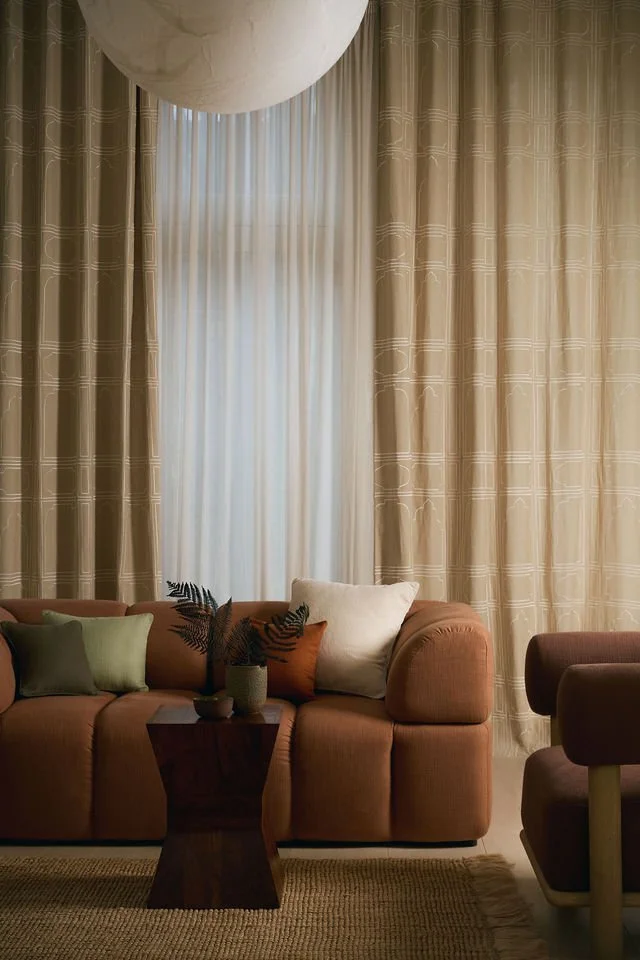 Living room with beige curtains, a puffed brown sofa with four cushions, a small dark wooden table with a potted plant, and another chair on the right.