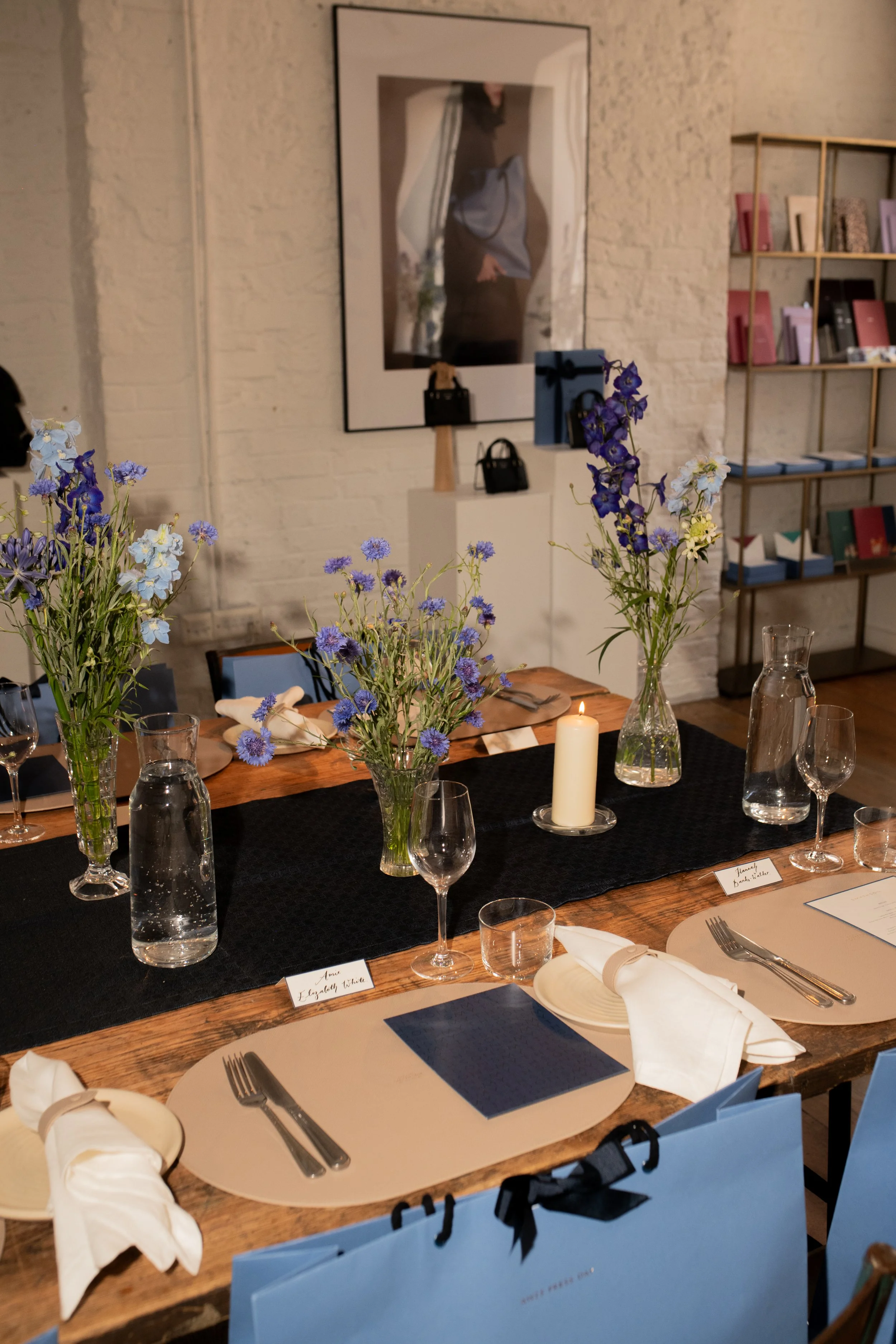 A decorated dining table with vases of blue and purple flowers, white napkins, candles, wine glasses, and water pitchers in a well-lit room. There are name tags and a gift bag on the table, with artwork and shelves in the background.
