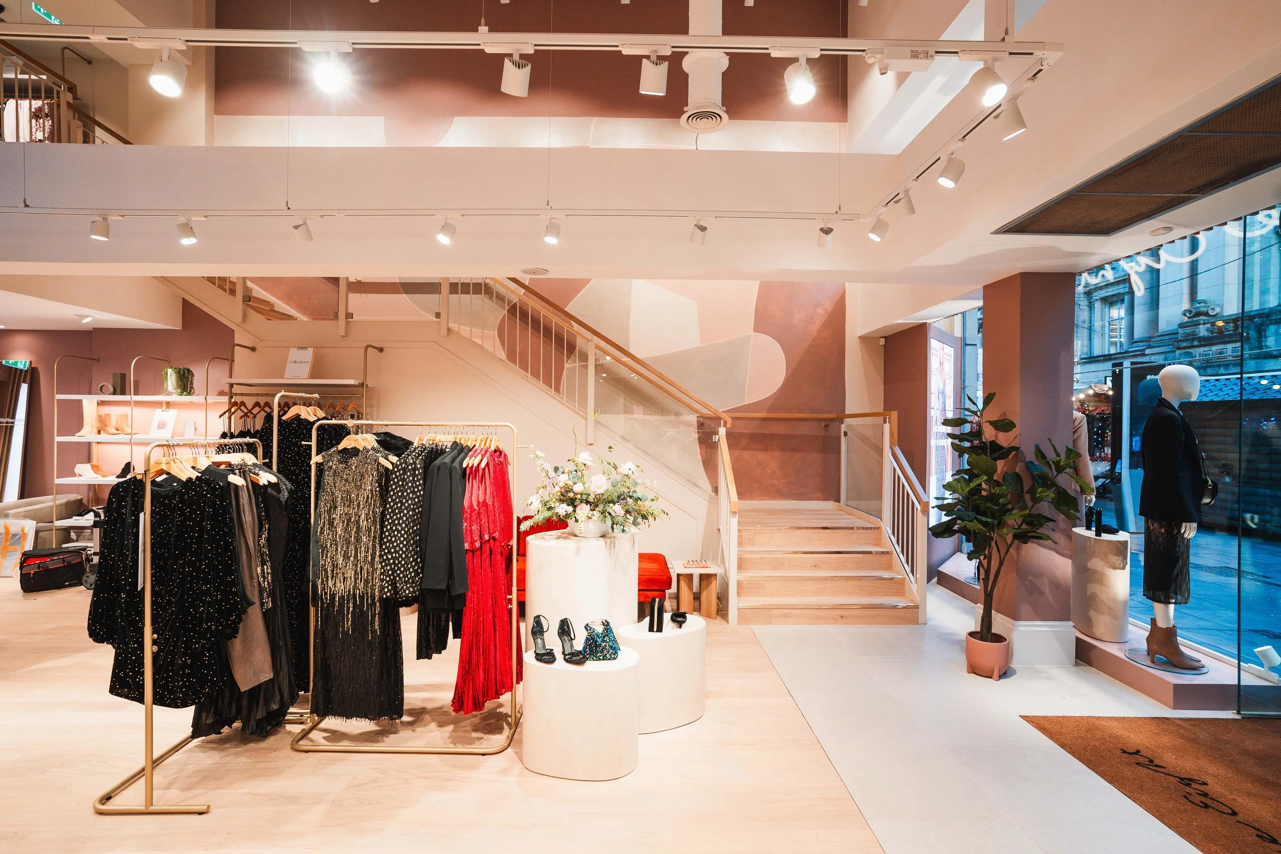 Interior of a boutique clothing store with clothing racks, a mannequin dressed in black, and a large window showing street outside.