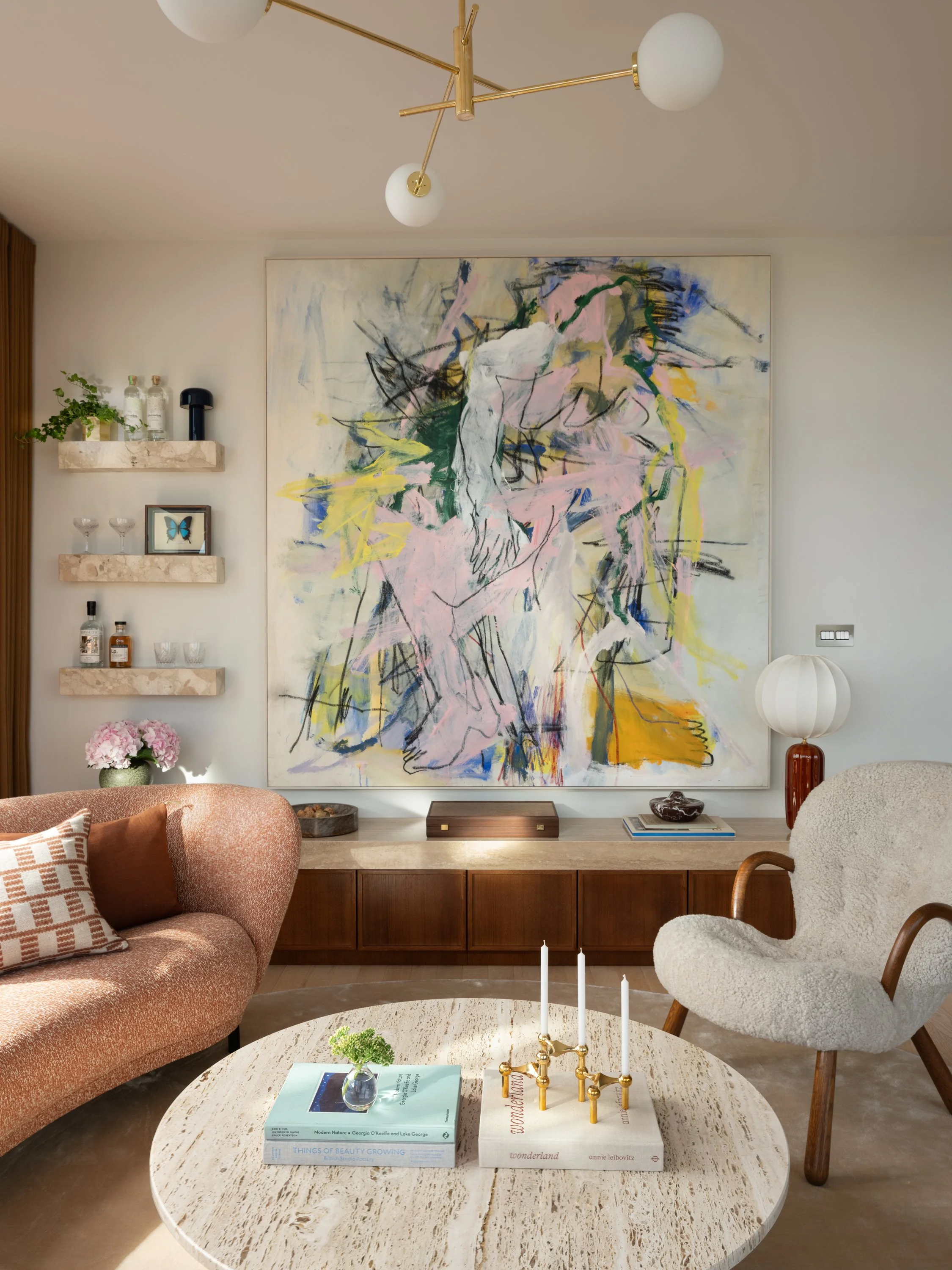 Living room with a large abstract painting, a peach couch, a white cozy armchair, a marble coffee table with books and candles, and a modern gold chandelier.