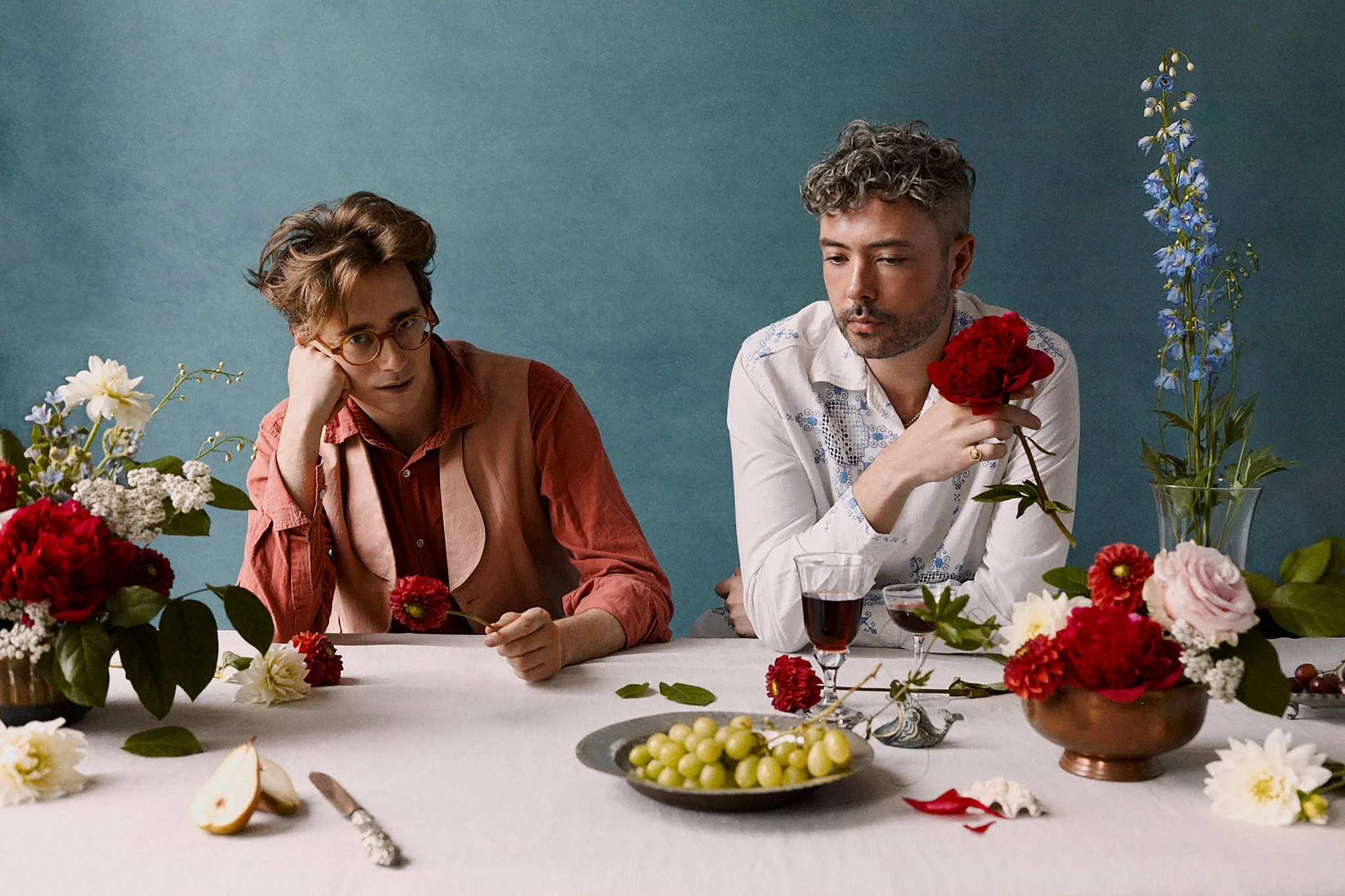 Two men sit at a table decorated with flowers, grapes, and wine. The man on the left has brown hair, glasses, and is wearing an orange shirt with a pink vest. The man on the right has curly gray hair, a beard, and is wearing a white embroidered shirt