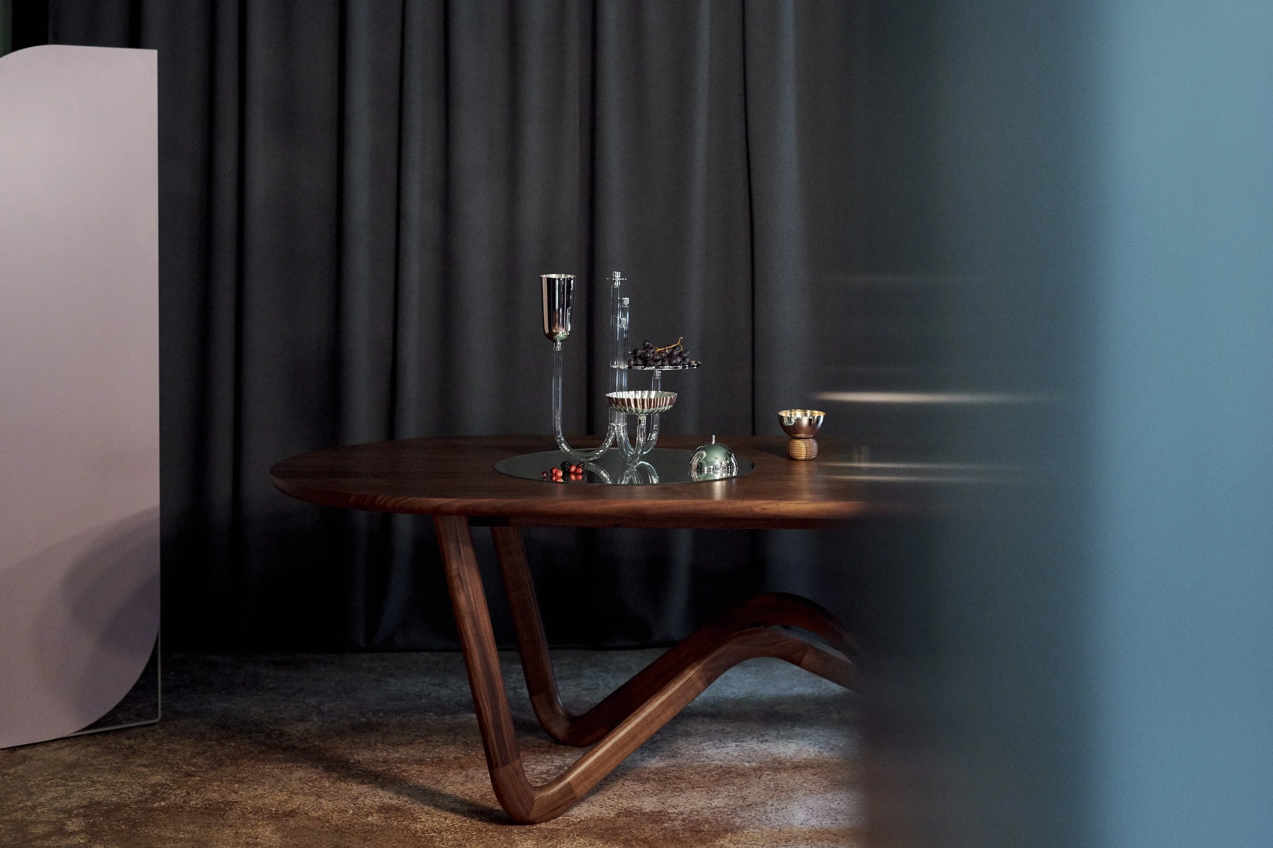 A wooden table with modern glassware, including a tall candlestick holder, a small glass bowl, and a small copper cup, set against a dark curtain backdrop.